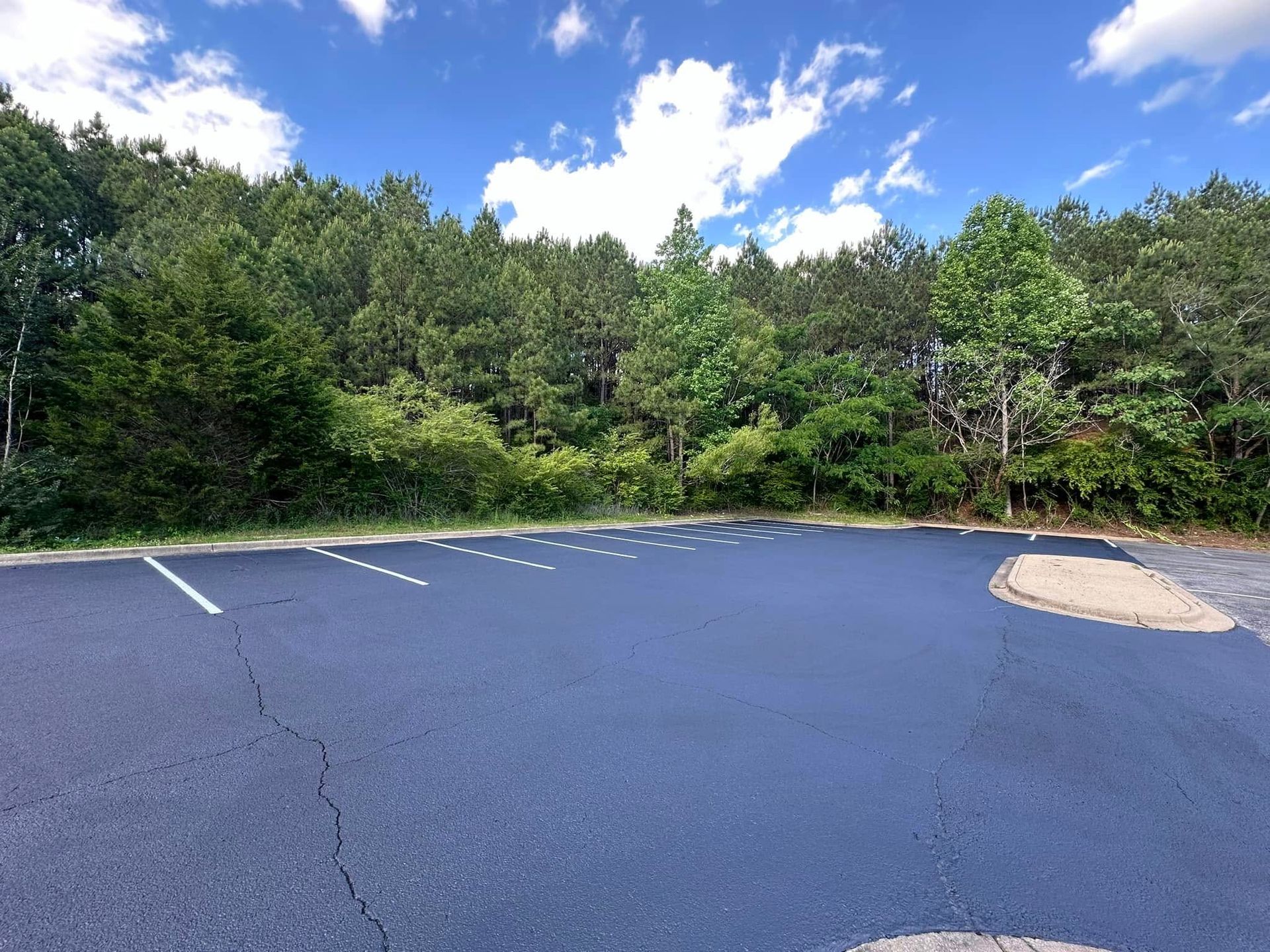 Empty paved parking lot with white parking space lines in front of a forest of green trees under a blue sky with clouds.
