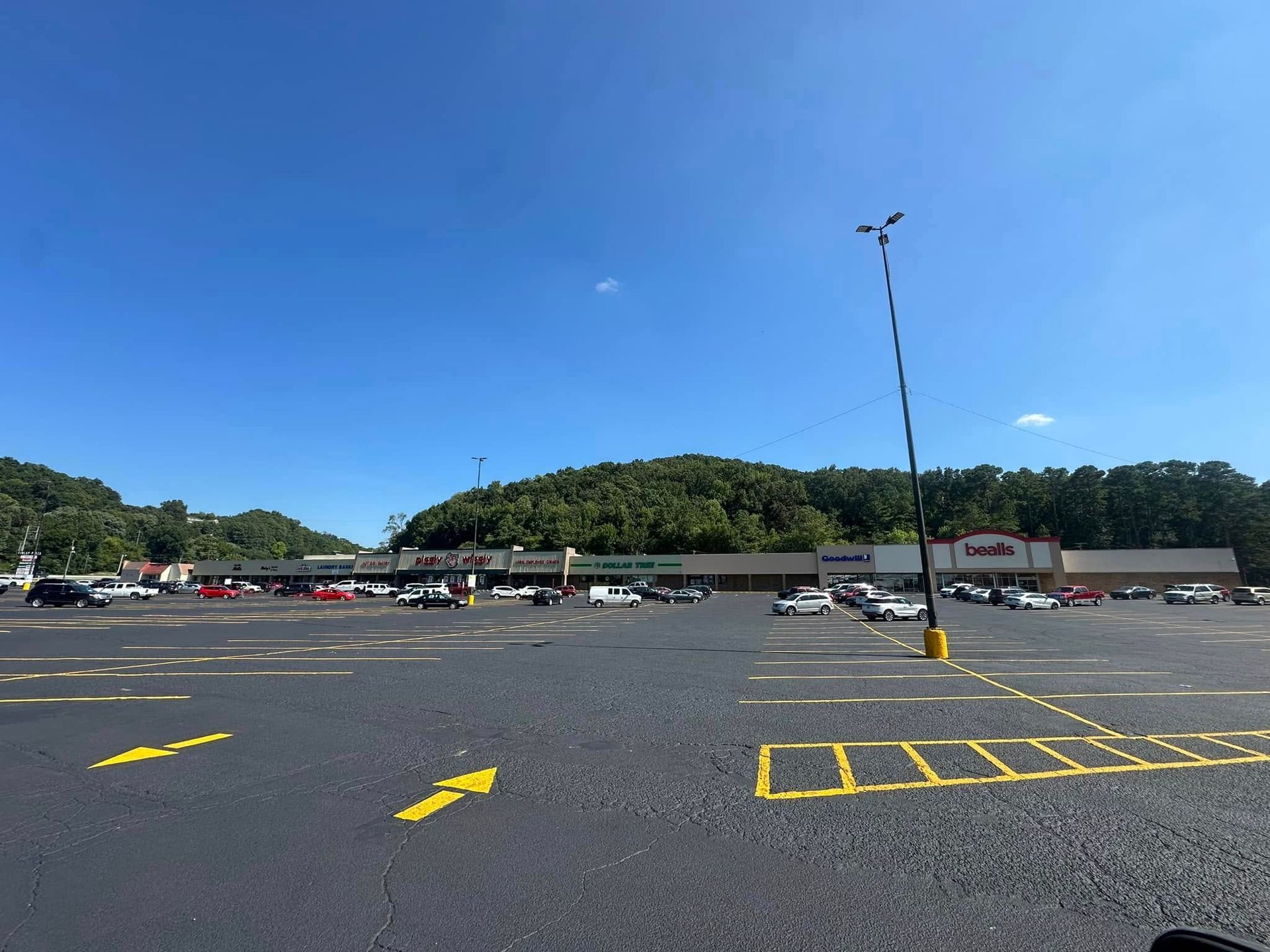 Exterior view of a shopping center parking lot on a sunny day. Cars are parked. Green trees are in the background.