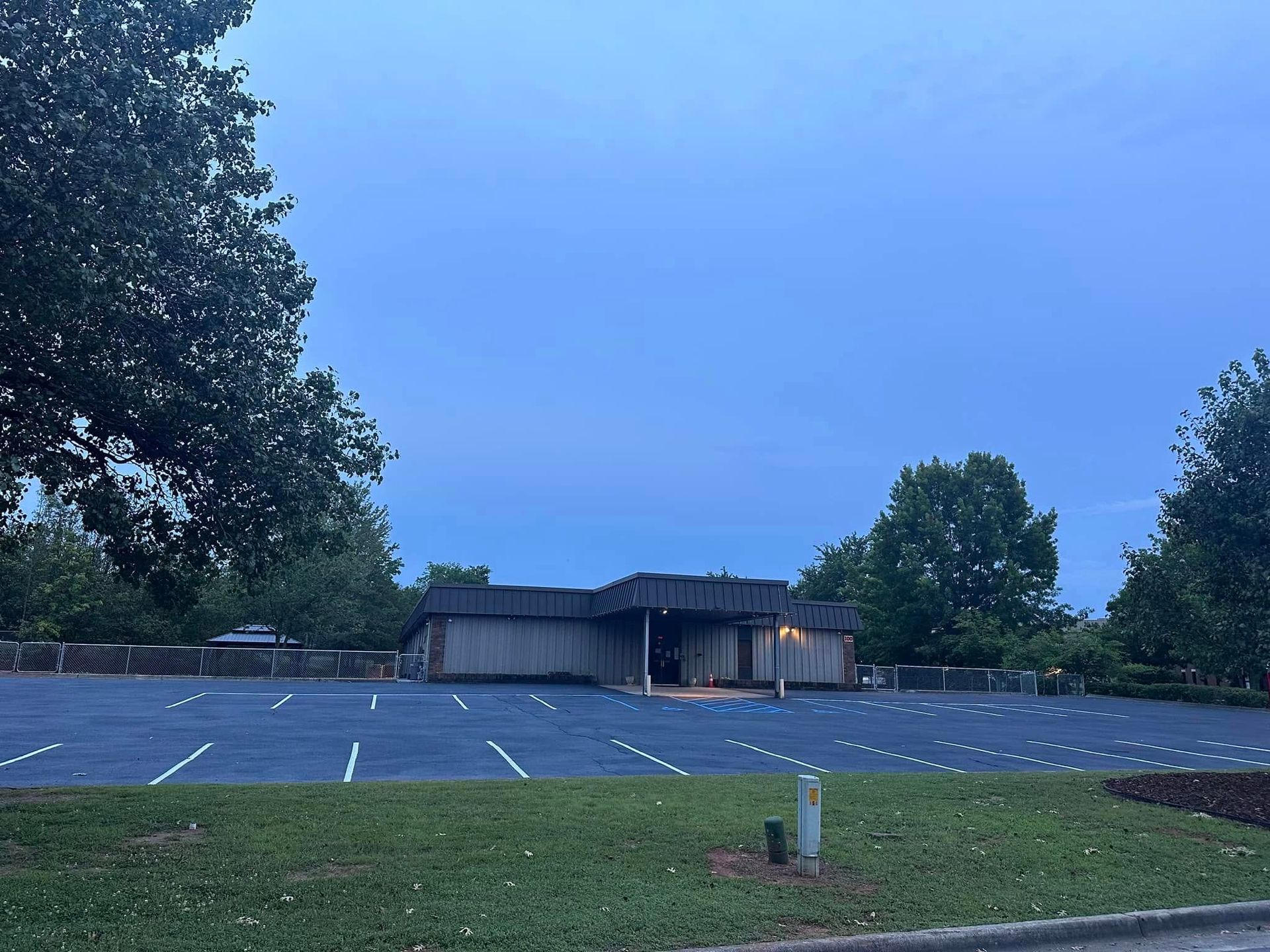 Empty parking lot in front of a low, wooden building under a blue sky. Trees frame the scene.