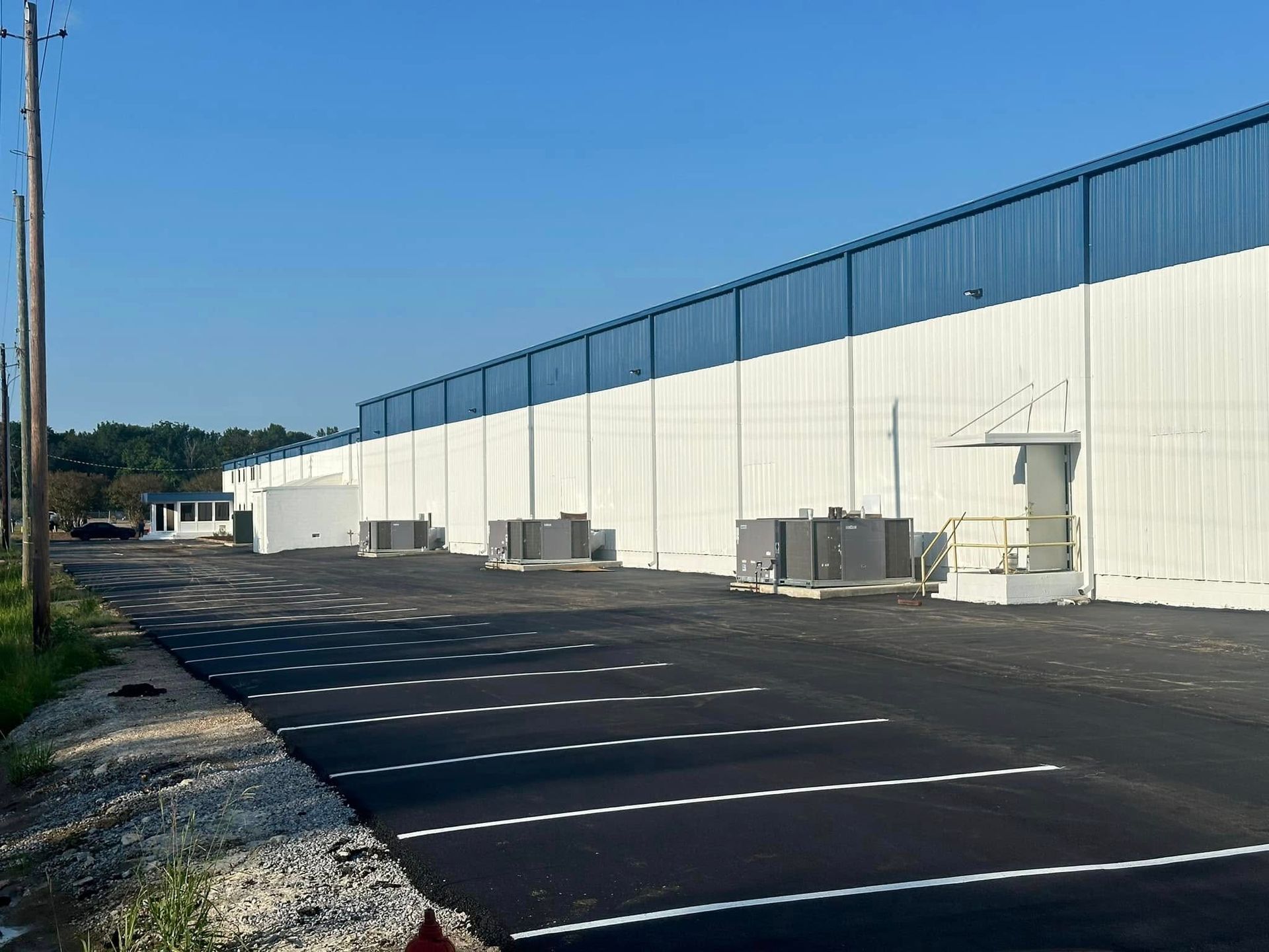 Large warehouse with blue and white exterior, parking lot in front, and utility poles alongside the road.