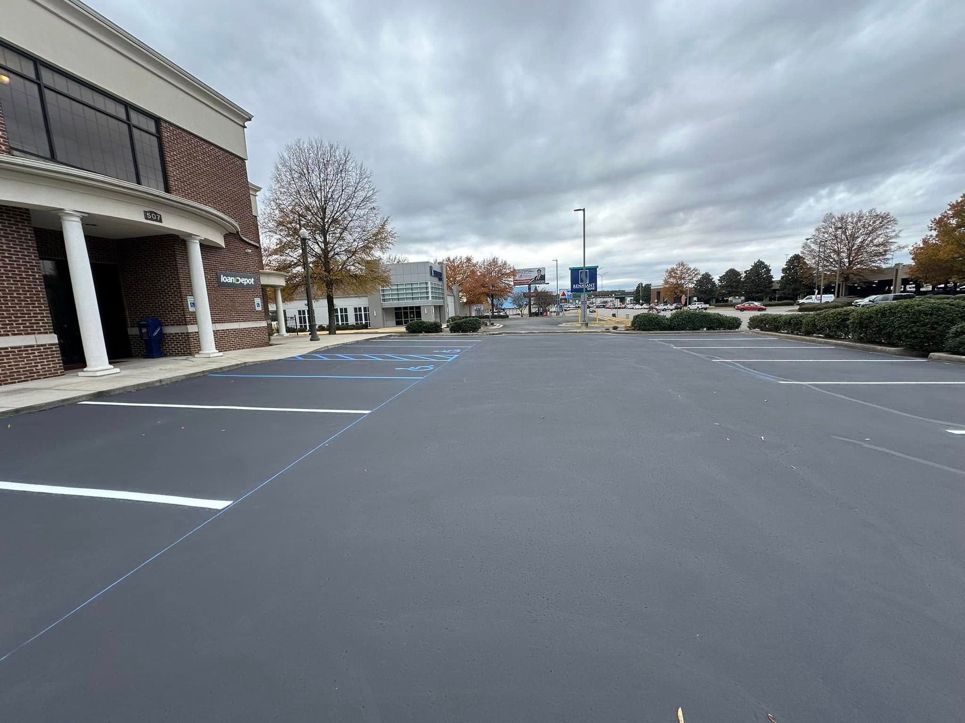 Newly paved, empty parking lot in front of a brick building and other businesses under a cloudy sky.