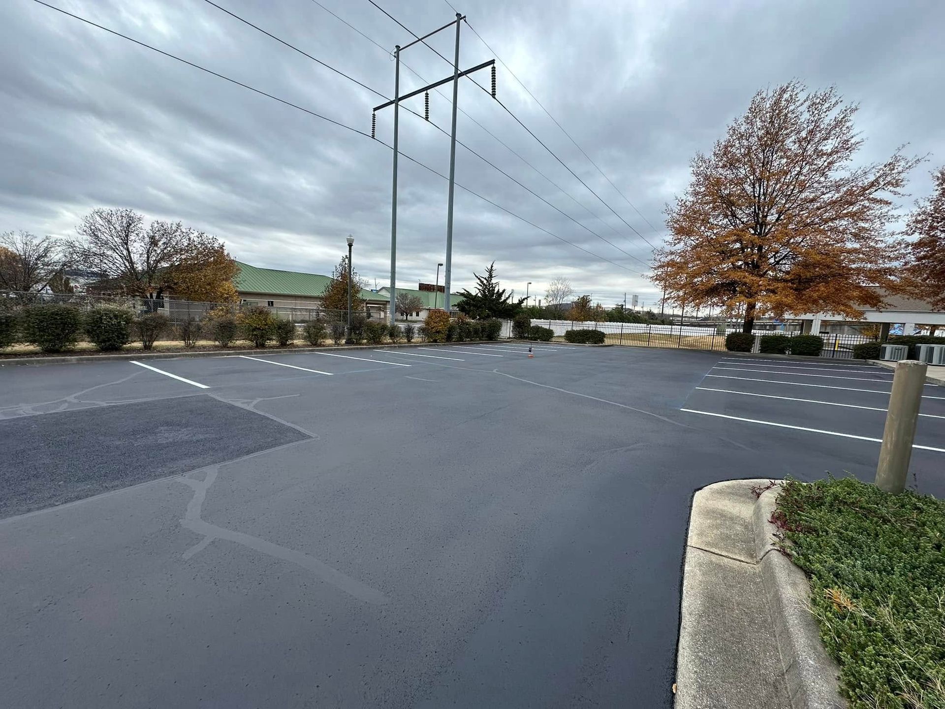 Empty asphalt parking lot under a cloudy sky, power lines overhead, with trees and a building in the background.