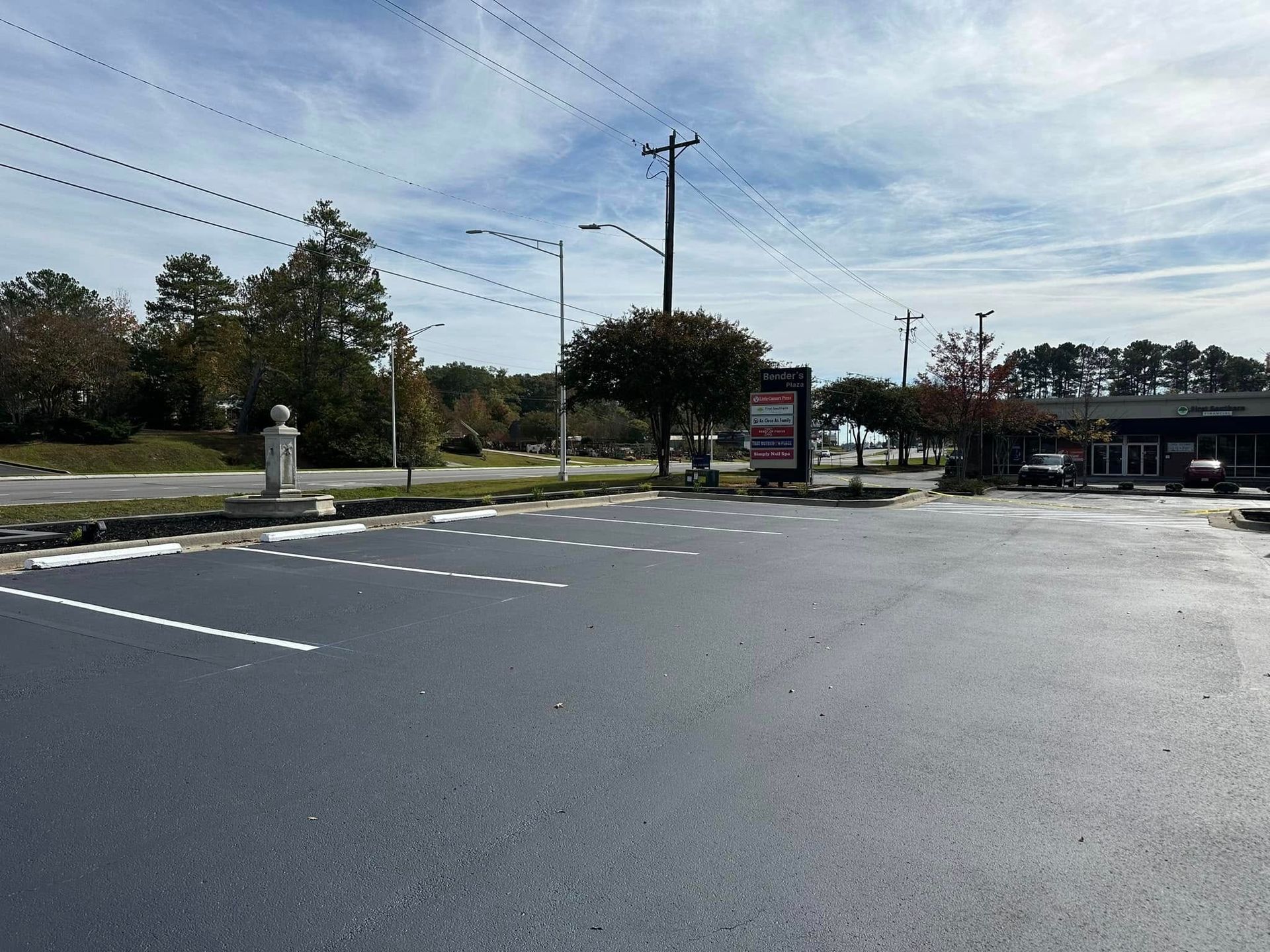 Newly paved asphalt parking lot on a sunny day with trees and power lines in background.