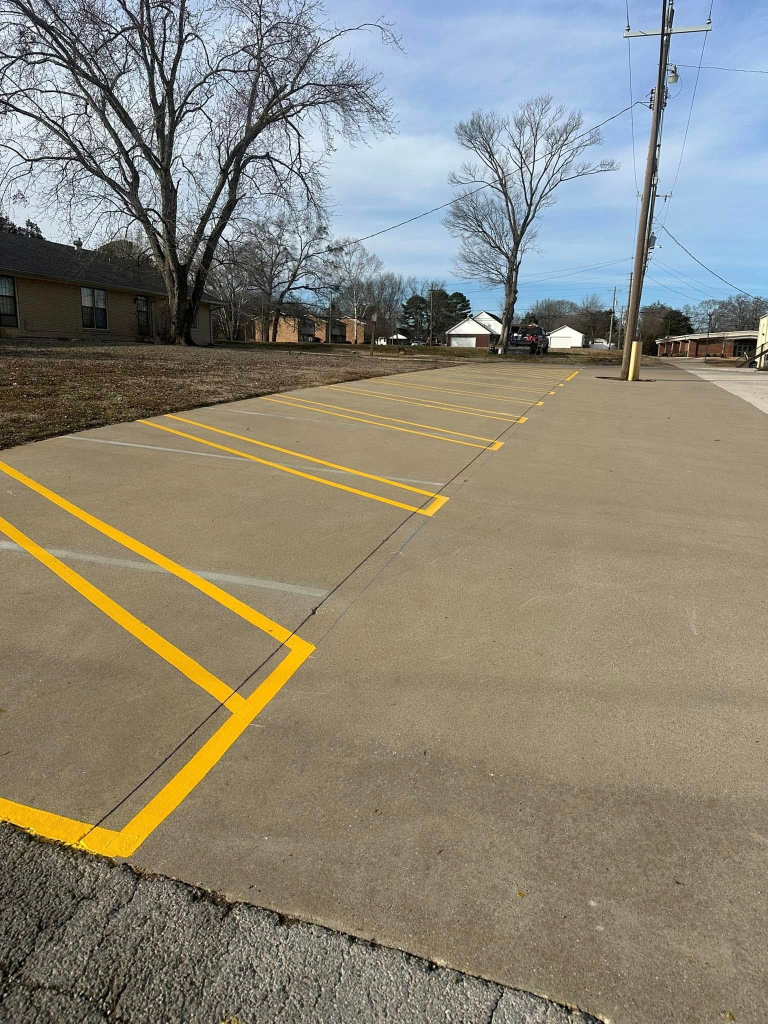 Concrete parking lot with yellow painted lines and open spaces. Bare trees and buildings in the background.