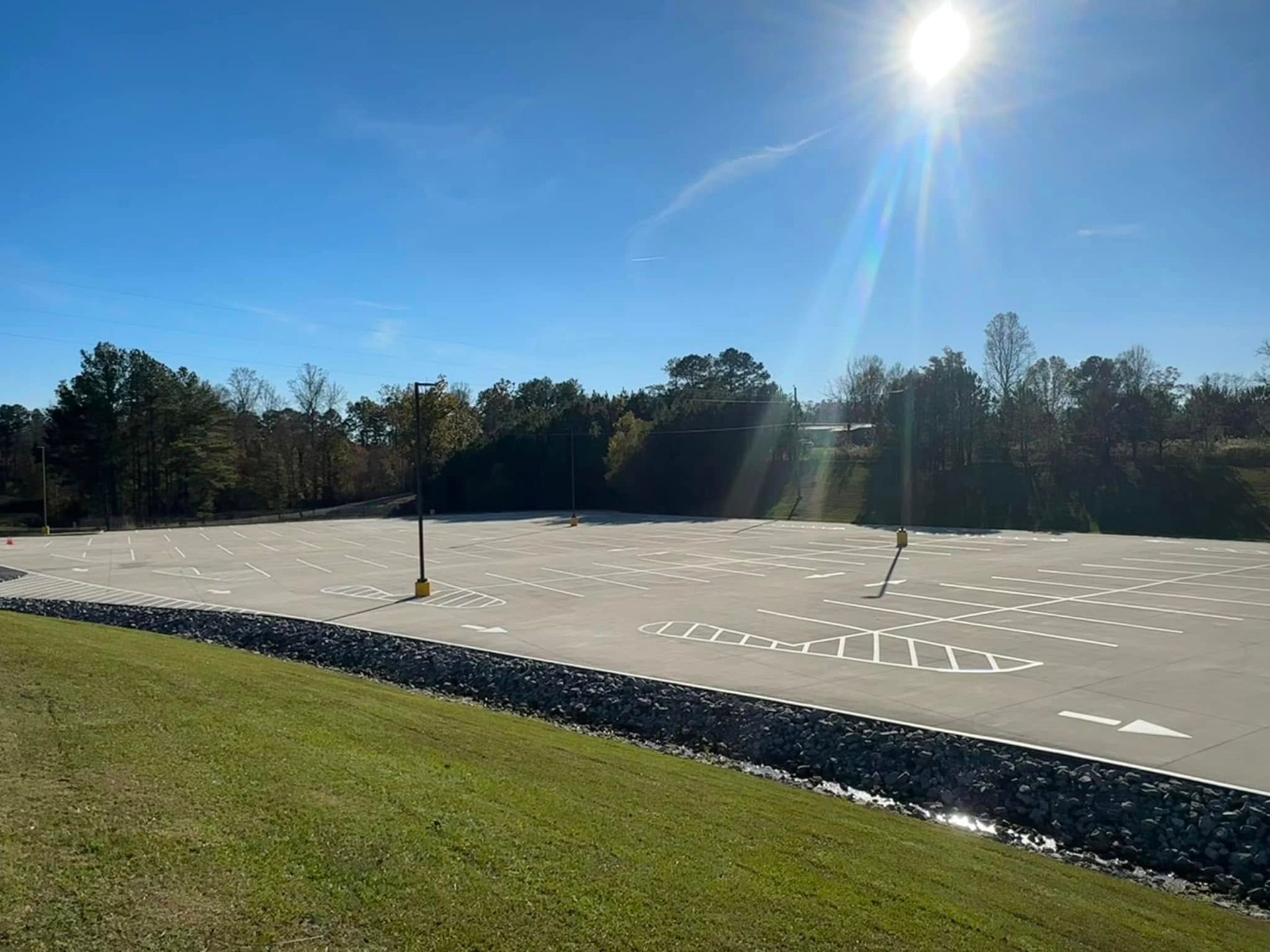 Empty parking lot with painted spaces, bordered by grass and trees under a sunny blue sky.