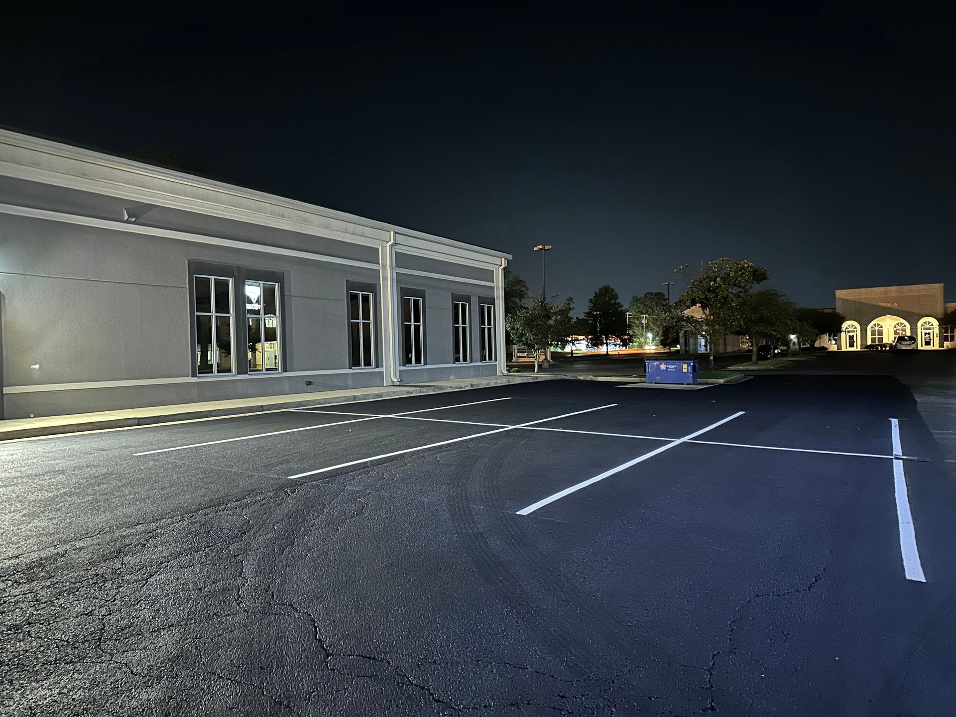 Nighttime exterior view of a gray building with windows and a freshly paved parking lot.