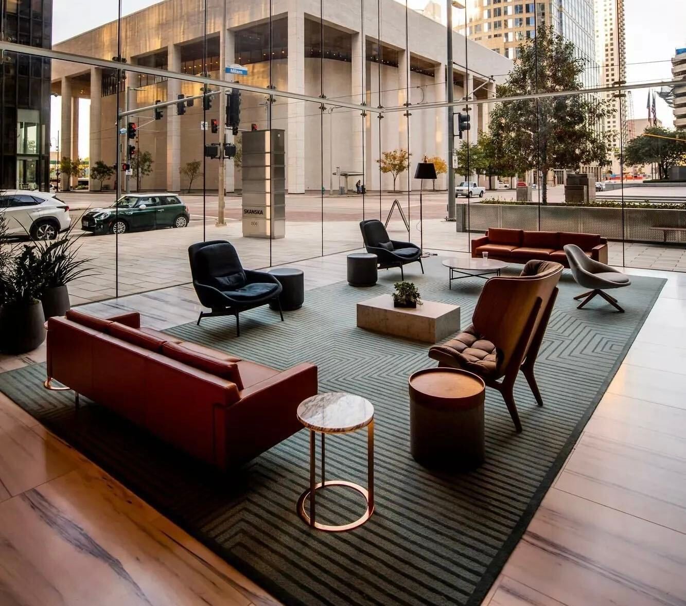 Modern lobby lounge with colorful chairs and tables on a woven rug beside floor-to-ceiling windows.