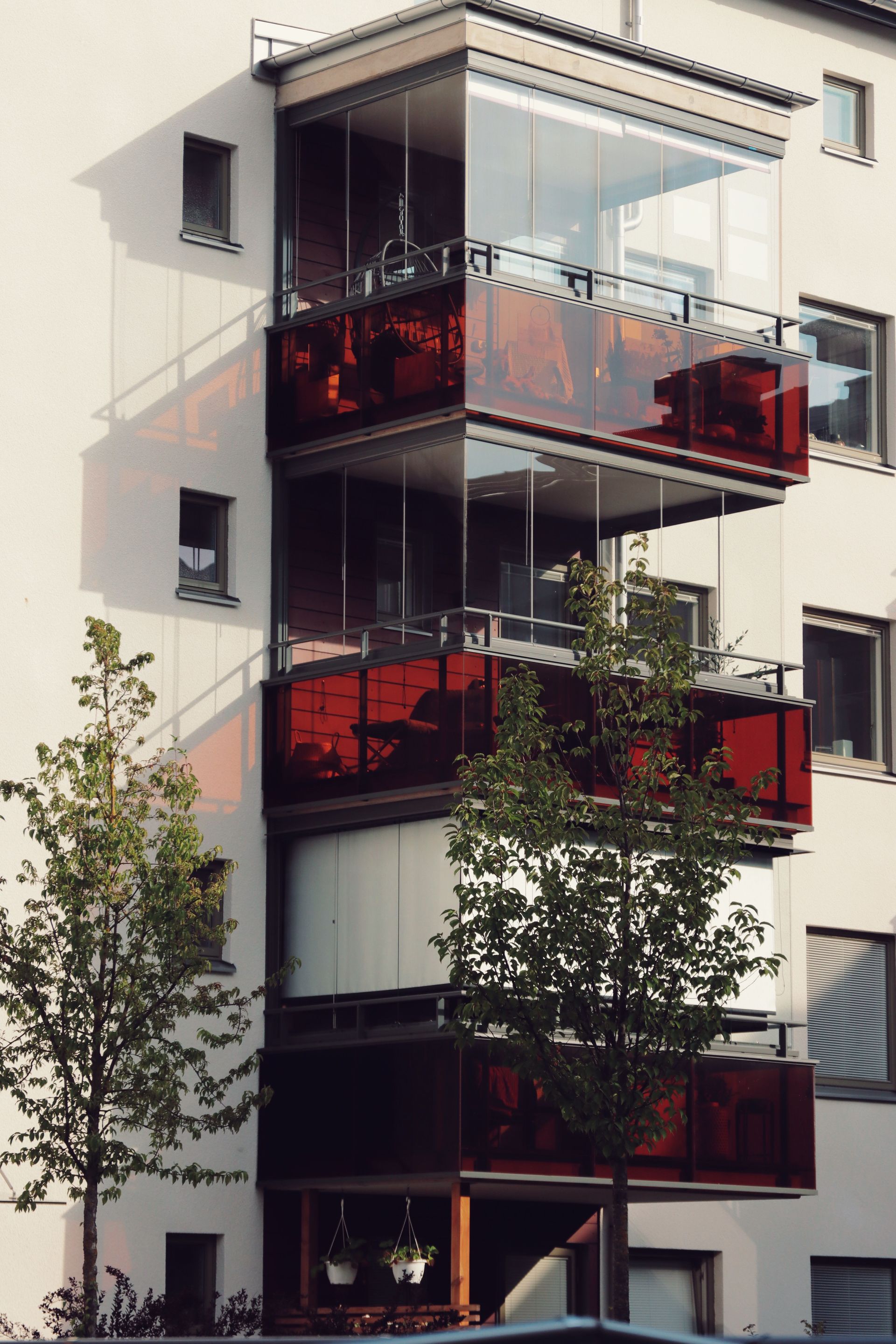Multi-story apartment building with glass balconies and red patio screens, partially hidden by trees