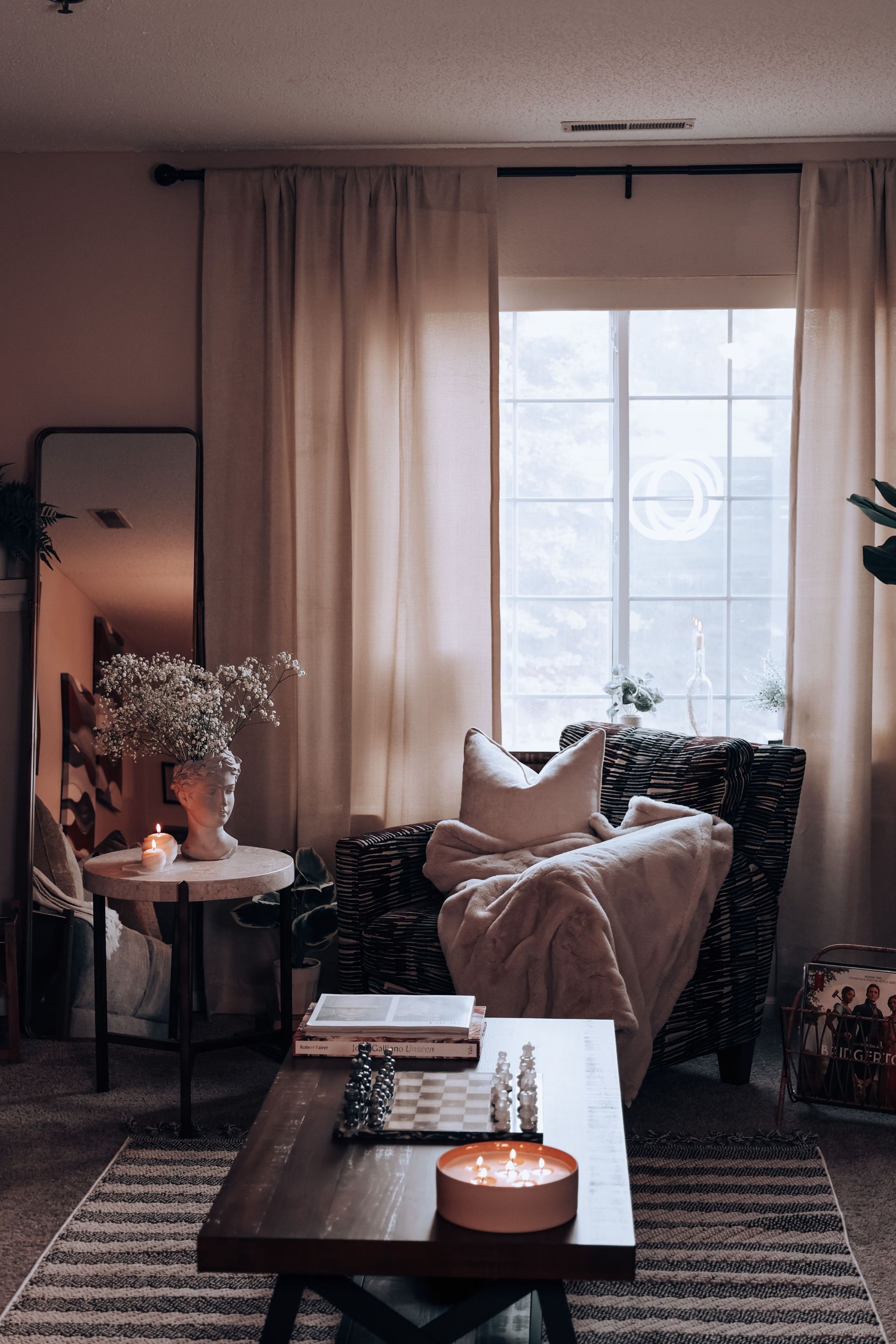 Cozy living room with a chair by a bright window, soft curtains, mirror, and a coffee table with books and a candle