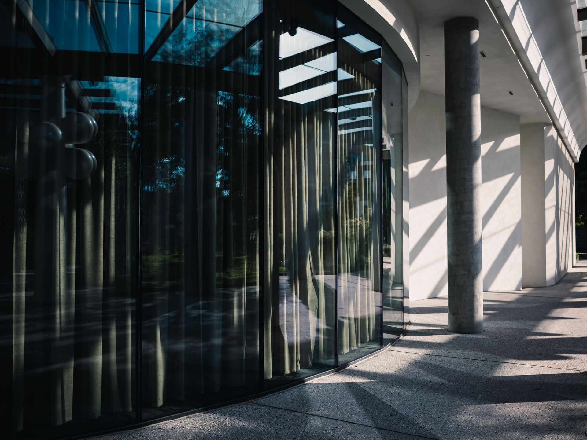 Modern glass building entrance with curved windows, striped shadows, and a bright walkway