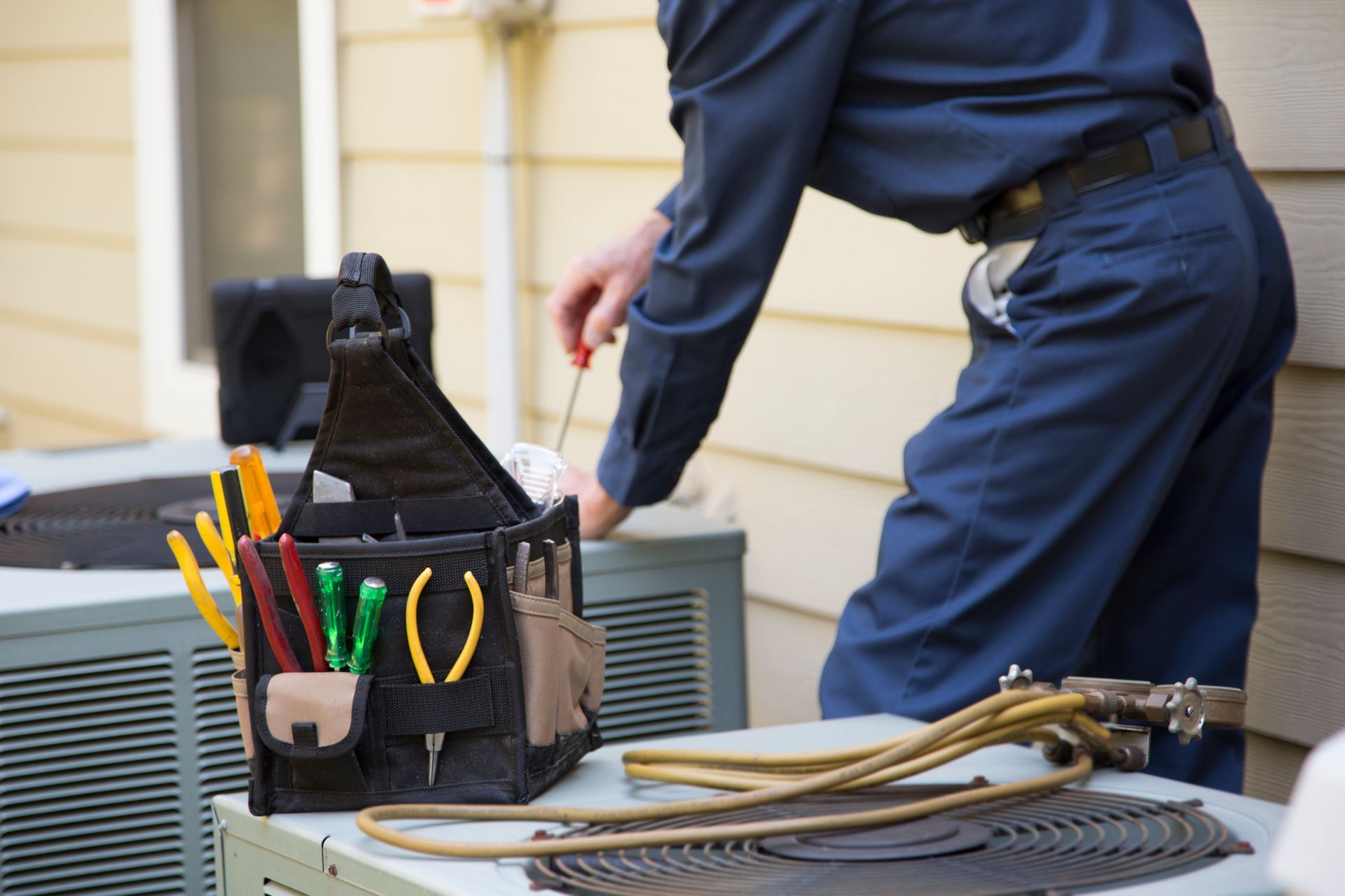 HVAC technician servicing outdoor AC unit with tools and hoses near house siding.
