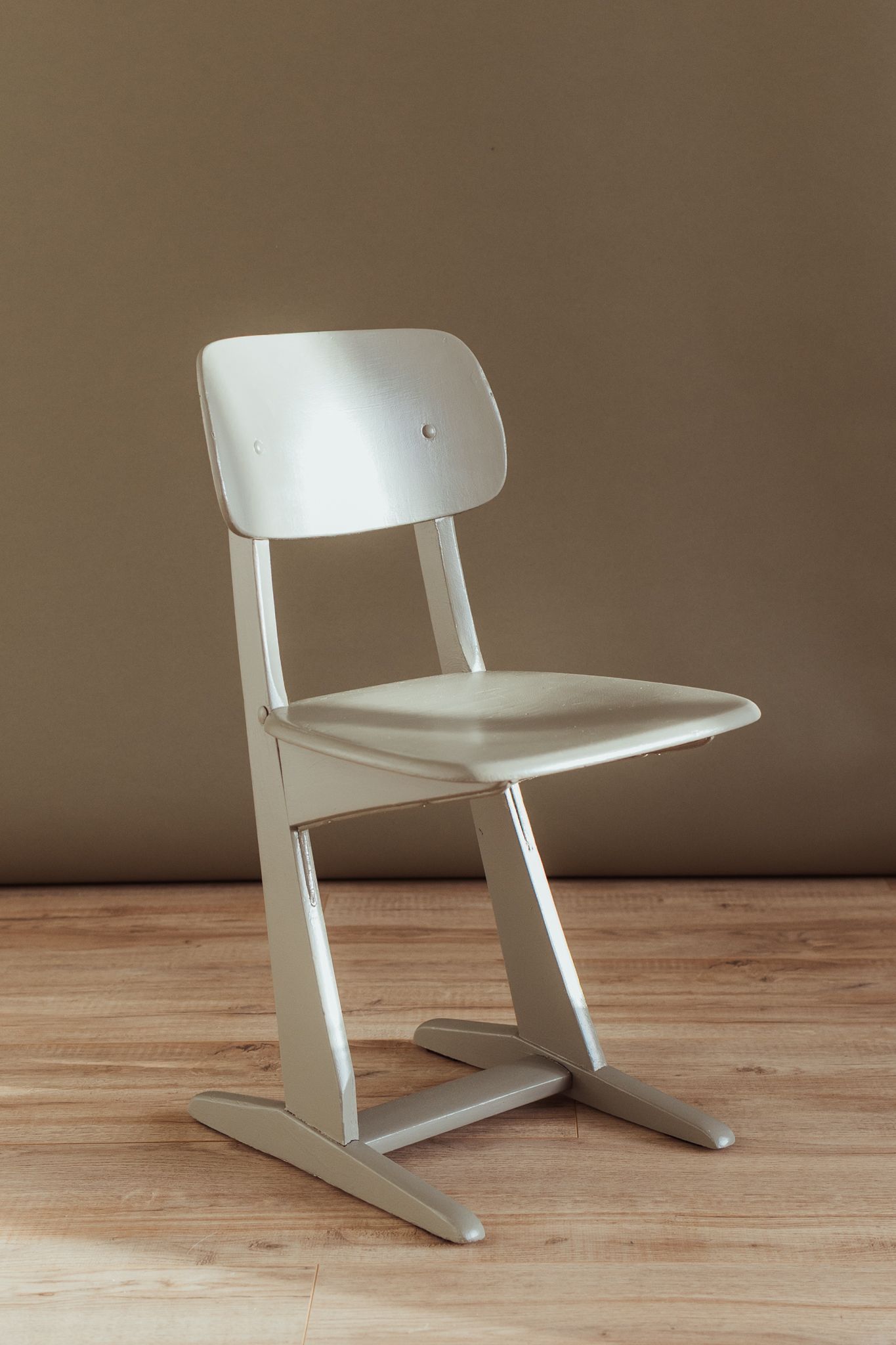 Een lichtgrijze, moderne houten stoel met een zwevend onderstel, geplaatst tegen een effen beige muur op een houten vloer.