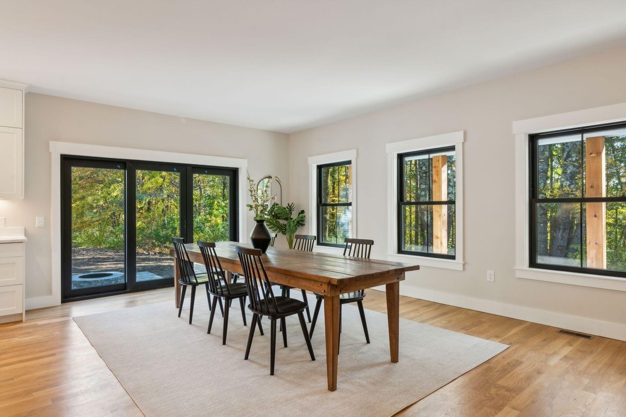 Bright dining room with wooden table, black chairs, large windows, and sliding glass doors overlooking greenery