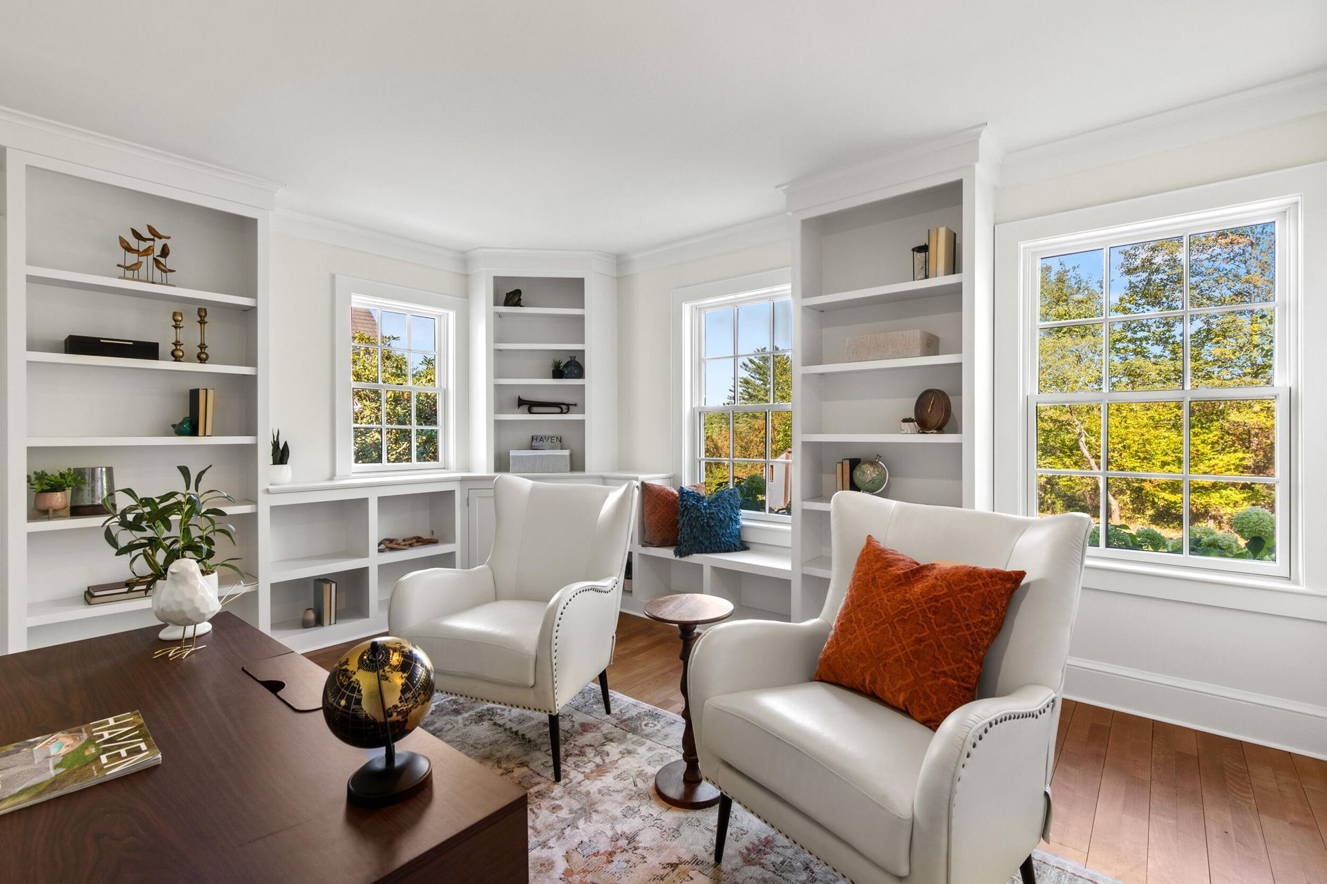 Bright living room with white built-in shelves, white chairs, wood floor, and orange accent pillows by sunny windows