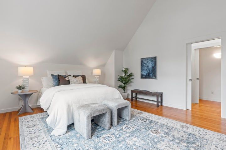 Bright bedroom with white bedding, two ottomans, wood floors, and a blue patterned rug.