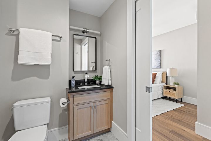 Small bathroom vanity with mirror and toilet, viewed through an open door into a bedroom.
