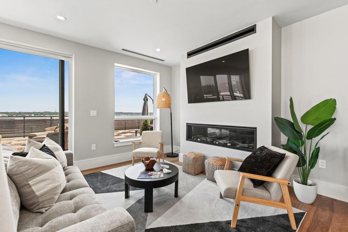 Modern living room with fireplace, wall-mounted TV, gray sofa, and large windows overlooking a deck