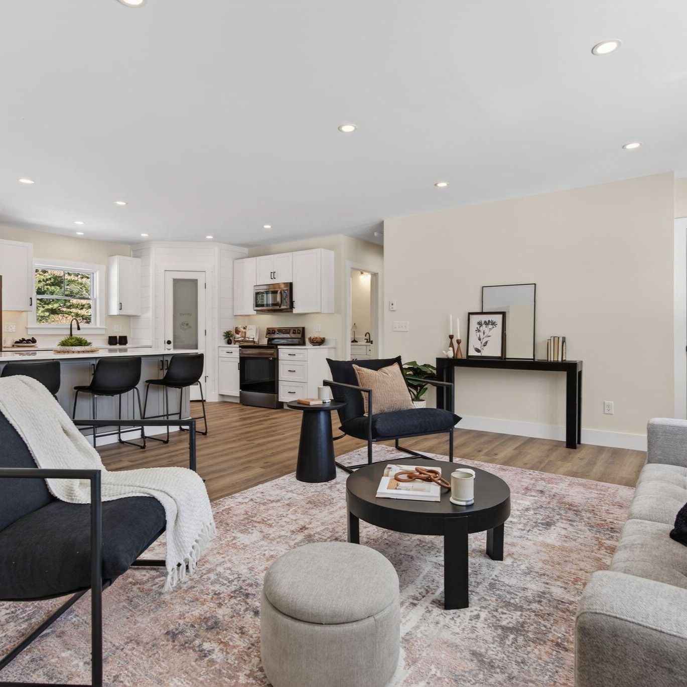 Bright open-concept living room with gray seating, black coffee table, patterned rug, and white kitchen in background
