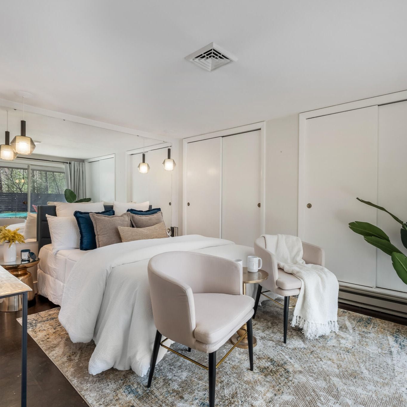 Bright modern bedroom with white bedding, beige chairs, blue pillows, and large built-in closets