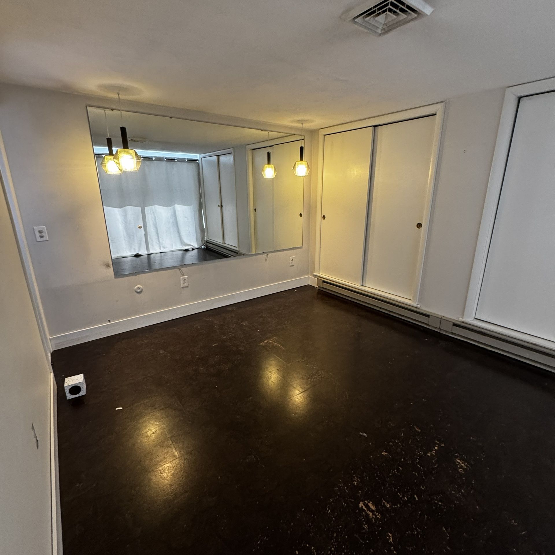 Empty room with dark wood floor, white walls, mirrors, and hanging pendant lights.