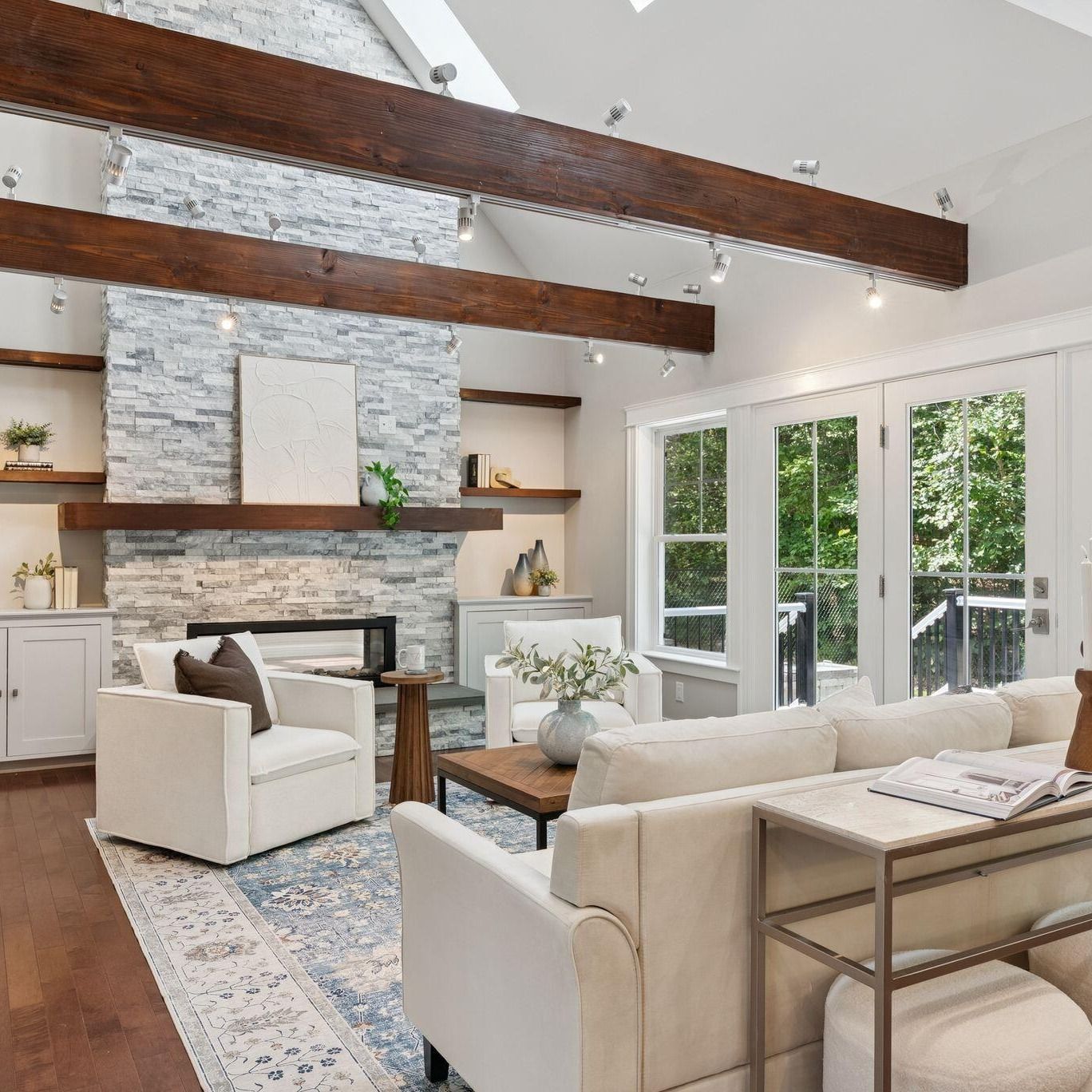 Bright living room with white sofa, stone fireplace, exposed beams, and French doors to a deck