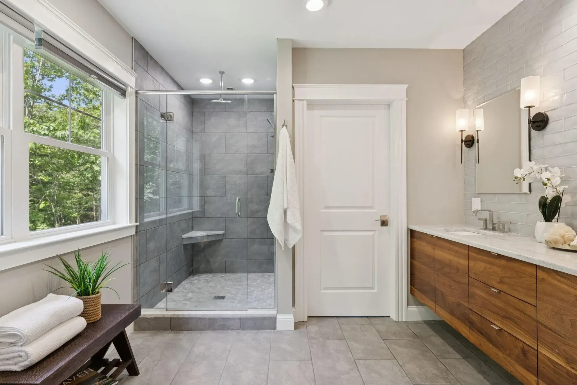 Bright modern bathroom with walk-in shower, window bench, vanity, and white door