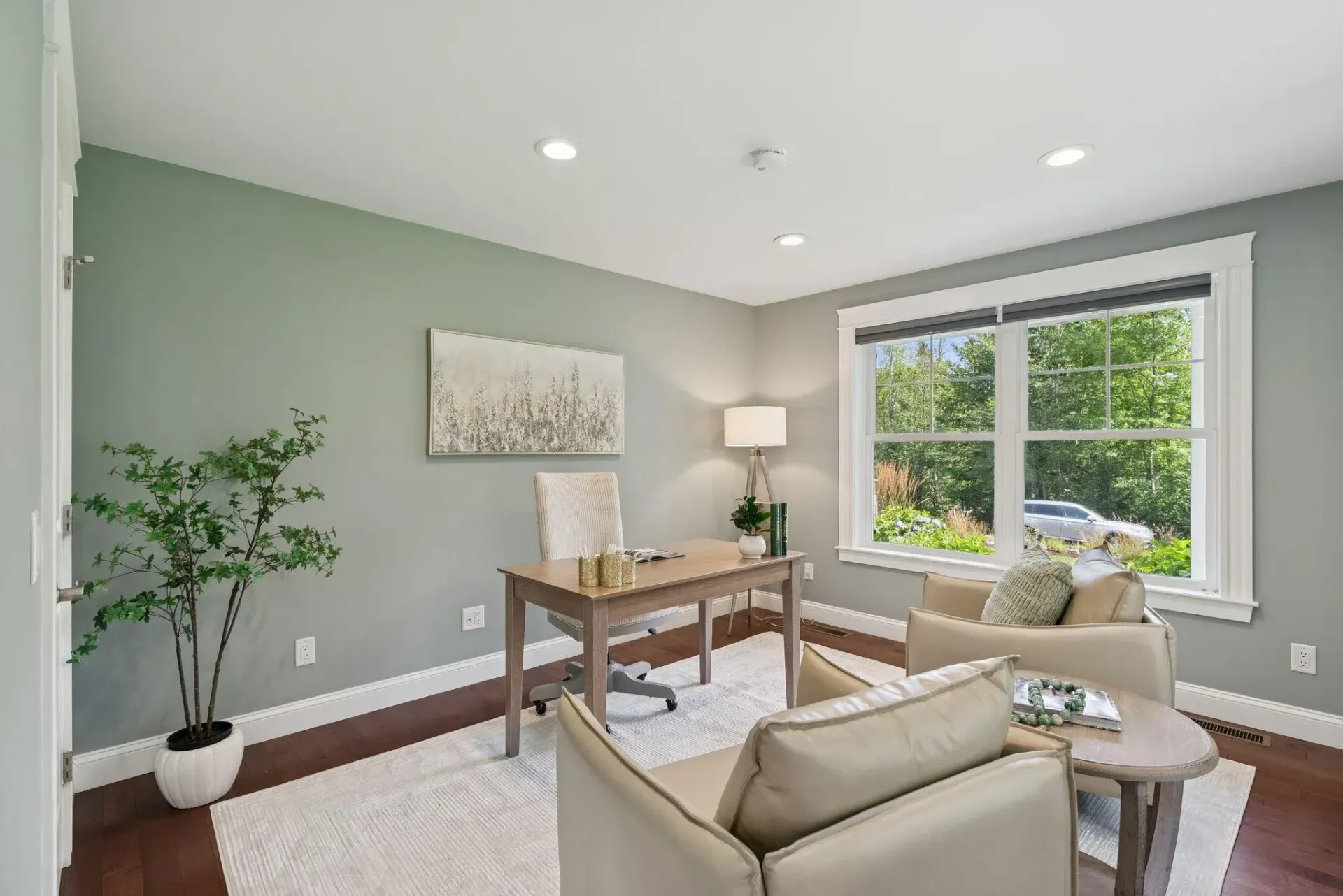 Bright living room with gray walls, beige chairs, desk, lamp, plant, and large window overlooking greenery