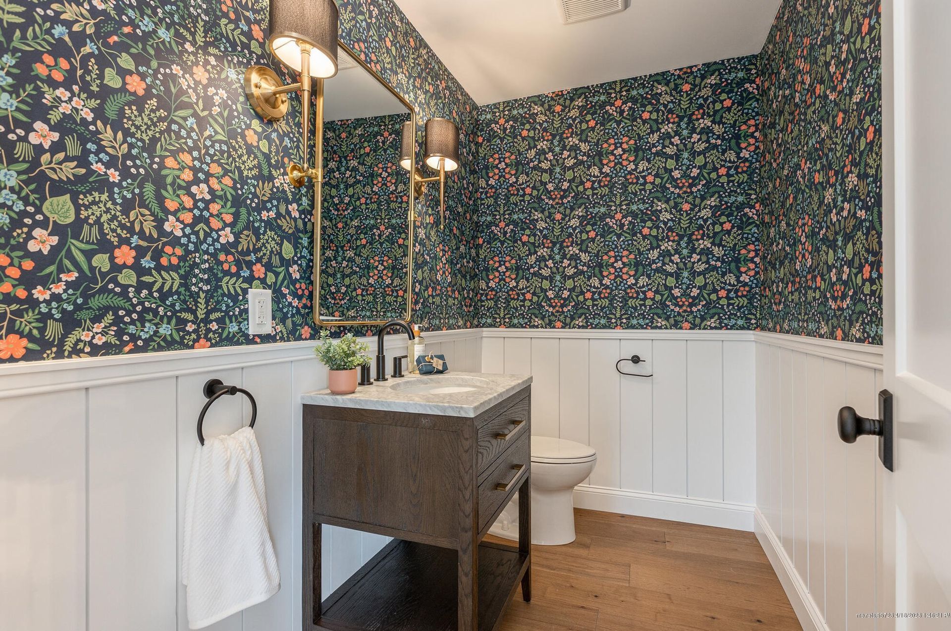 Bright bathroom with floral wallpaper, white wainscoting, wood vanity, sink, mirror, and toilet.