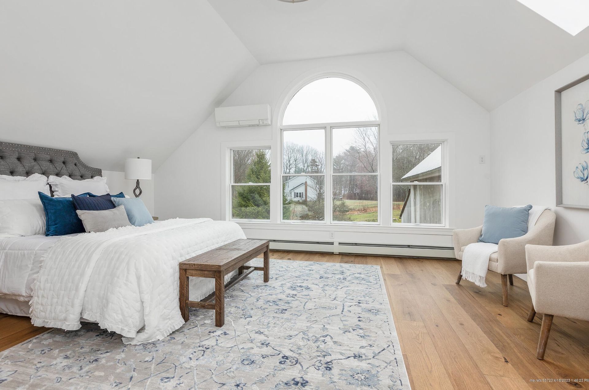 Bright bedroom with vaulted ceiling, arched window, white bed, blue accents, and beige chair on hardwood floor