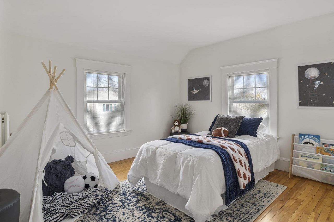 Bright bedroom with a white bed, teepee tent, stuffed animals, and patterned rug near two windows.