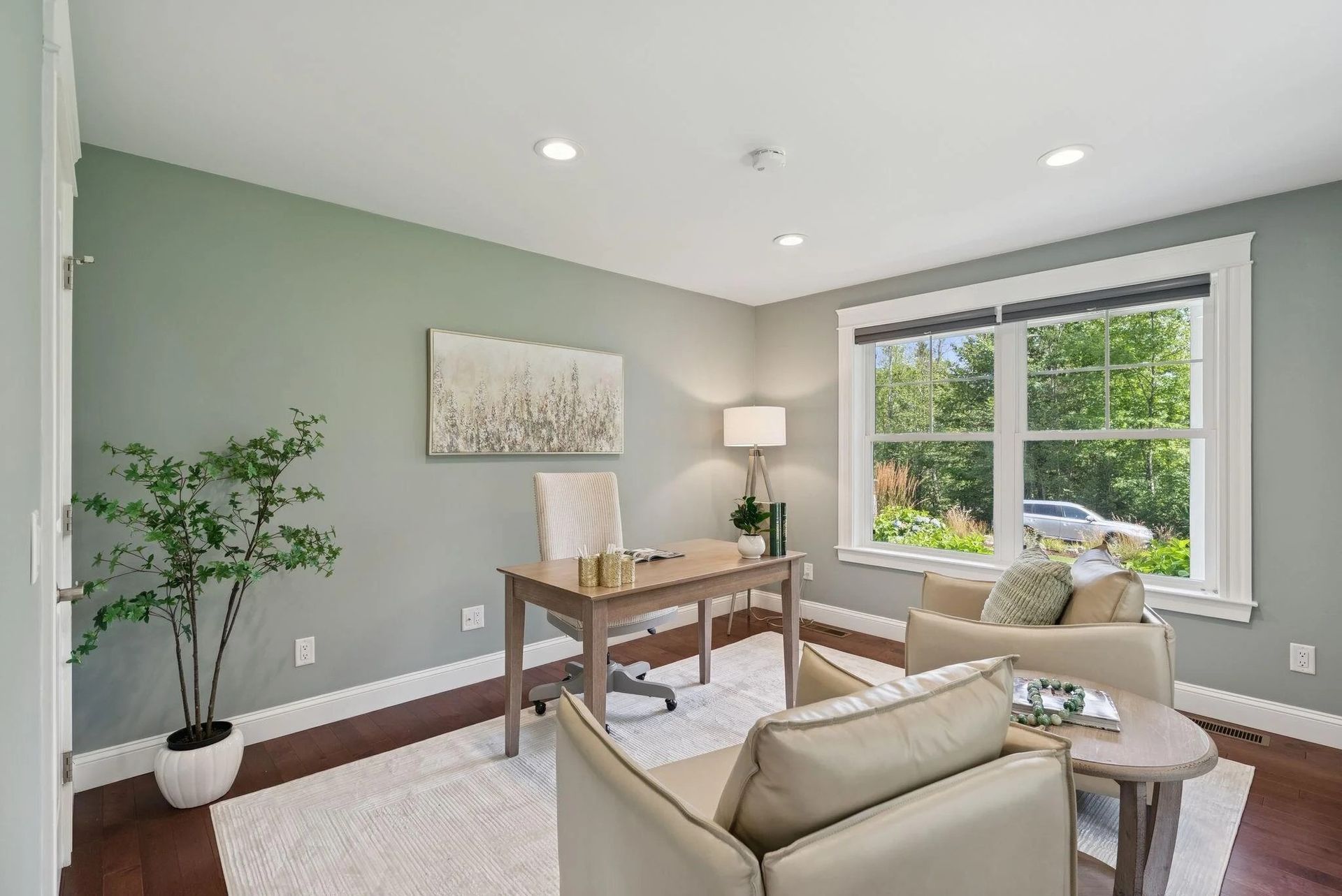 Bright living room with beige chairs, wooden table, green walls, and a large window overlooking trees