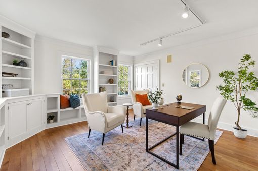 Bright living room with white built-ins, large windows, wooden desk, beige chairs, and potted plants