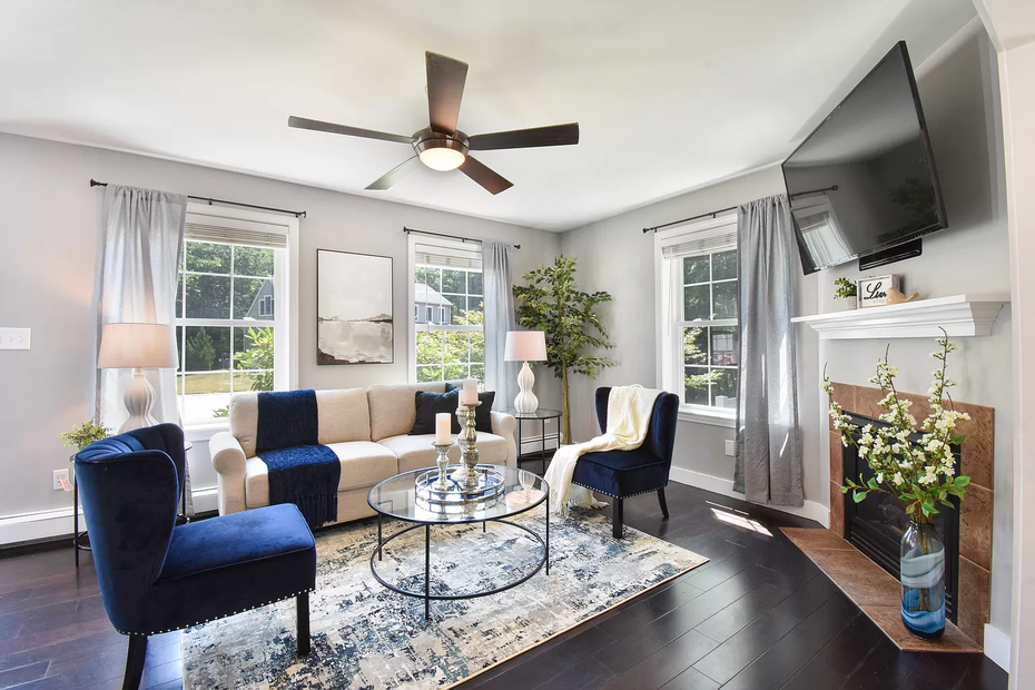 Bright living room with blue chairs, beige sofa, glass coffee table, fireplace, and wall-mounted TV.