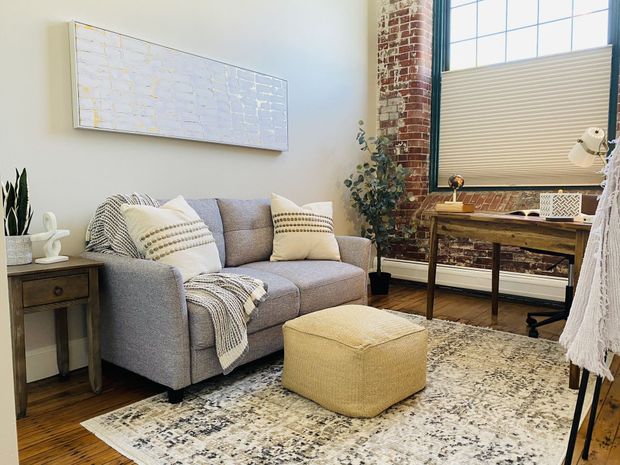 Cozy living room with gray loveseat, beige ottoman, patterned rug, and bright window by a desk