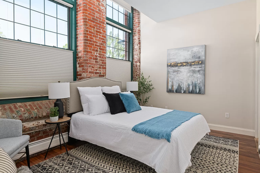 Bright bedroom with a made bed, large windows, exposed brick, and a gray chair.