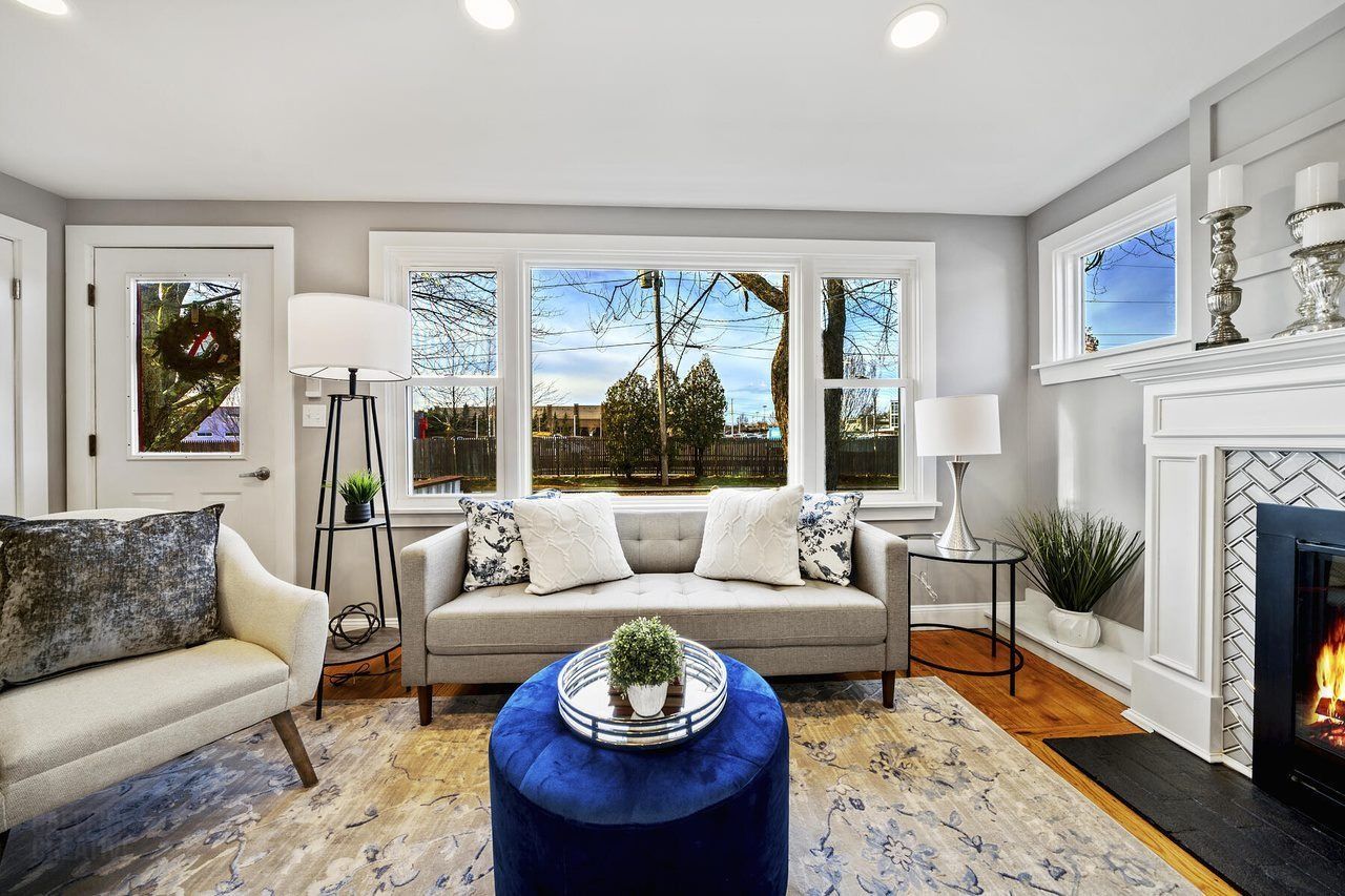 Bright living room with large windows, gray sofa, white armchairs, blue ottoman, and fireplace.