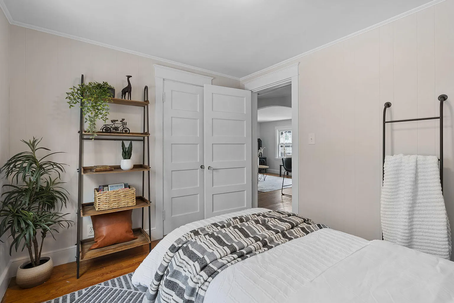 Bright bedroom with white walls, open double doors, bed, plant, shelf, and towel rack