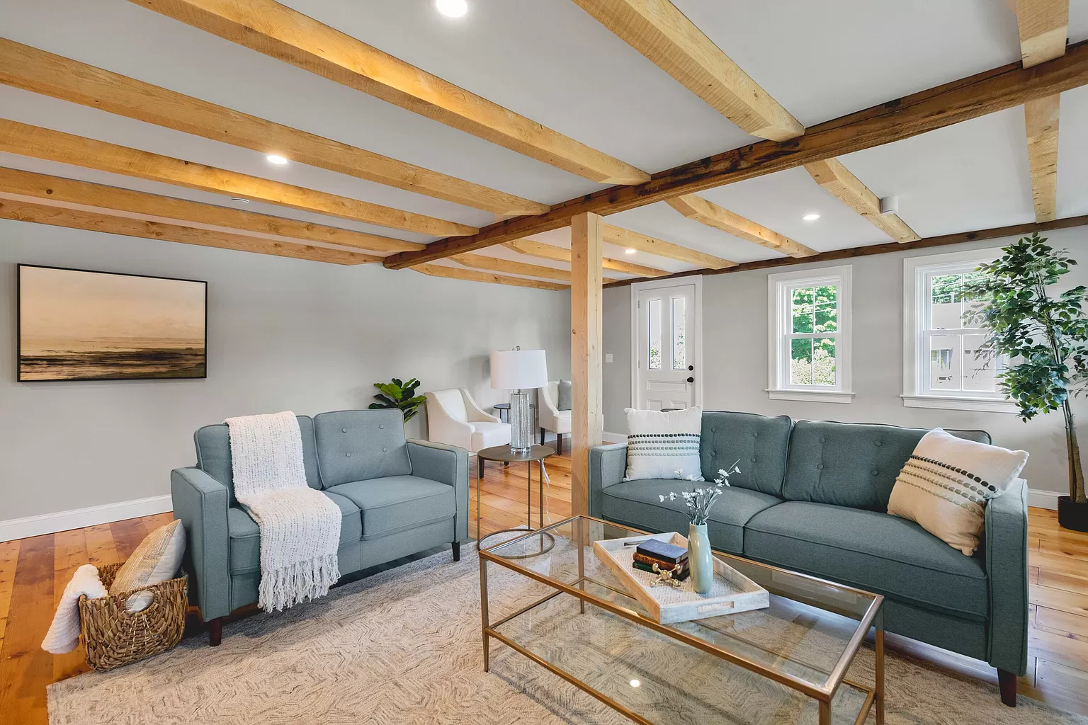 Bright living room with exposed wood beams, teal sofa, armchair, coffee table, and a potted plant.