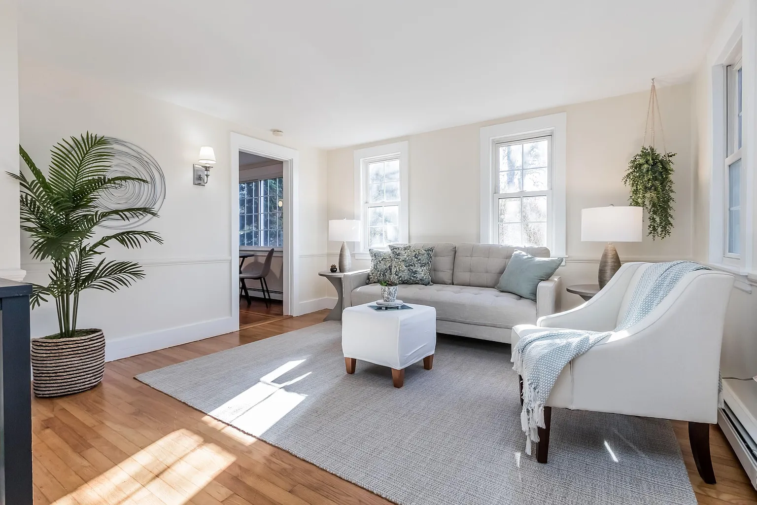 Bright living room with gray sofa, white armchair, potted plant, and sunlight on a rug.