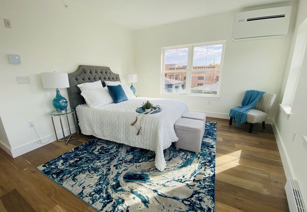 Bright bedroom with white bed, blue accents, patterned rug, and two windows overlooking greenery