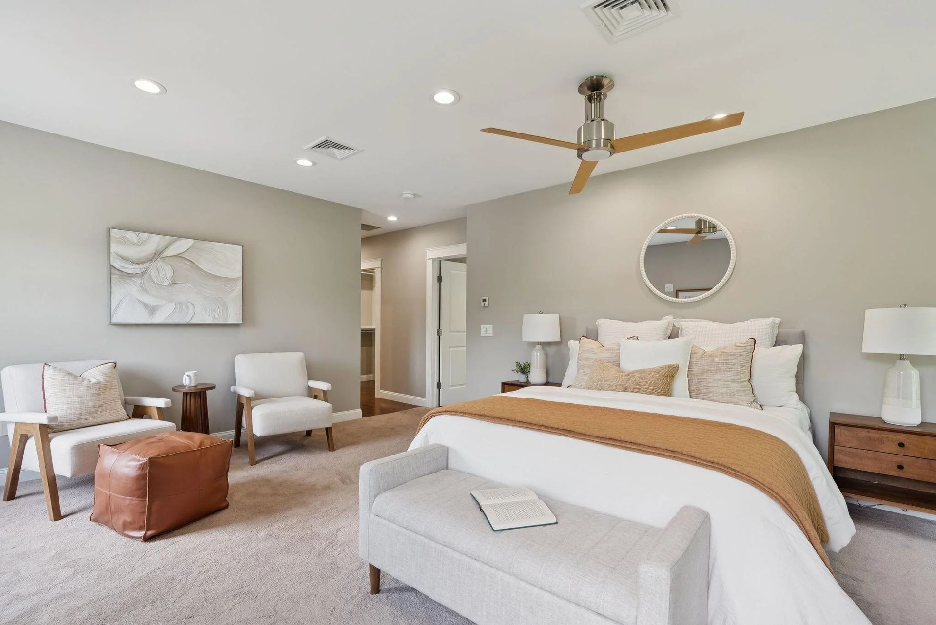 Bright, neutral bedroom with a bed, bench, two chairs, and a ceiling fan next to a sitting area