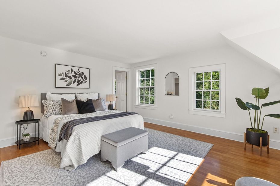 Bright bedroom with a bed, gray bench, hardwood floors, and sunlight through two windows