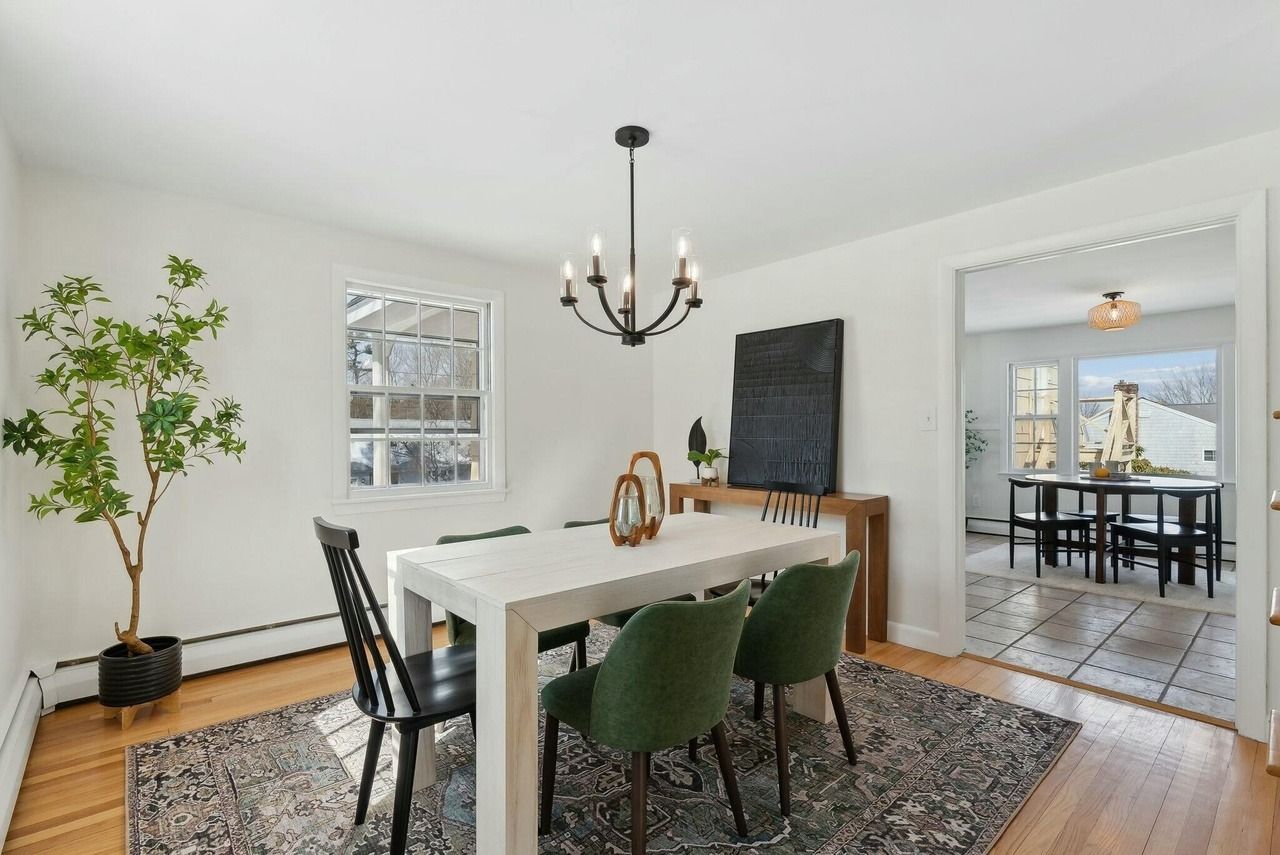 Bright dining room with white table, green chairs, chandelier, plant, and open view into a second room