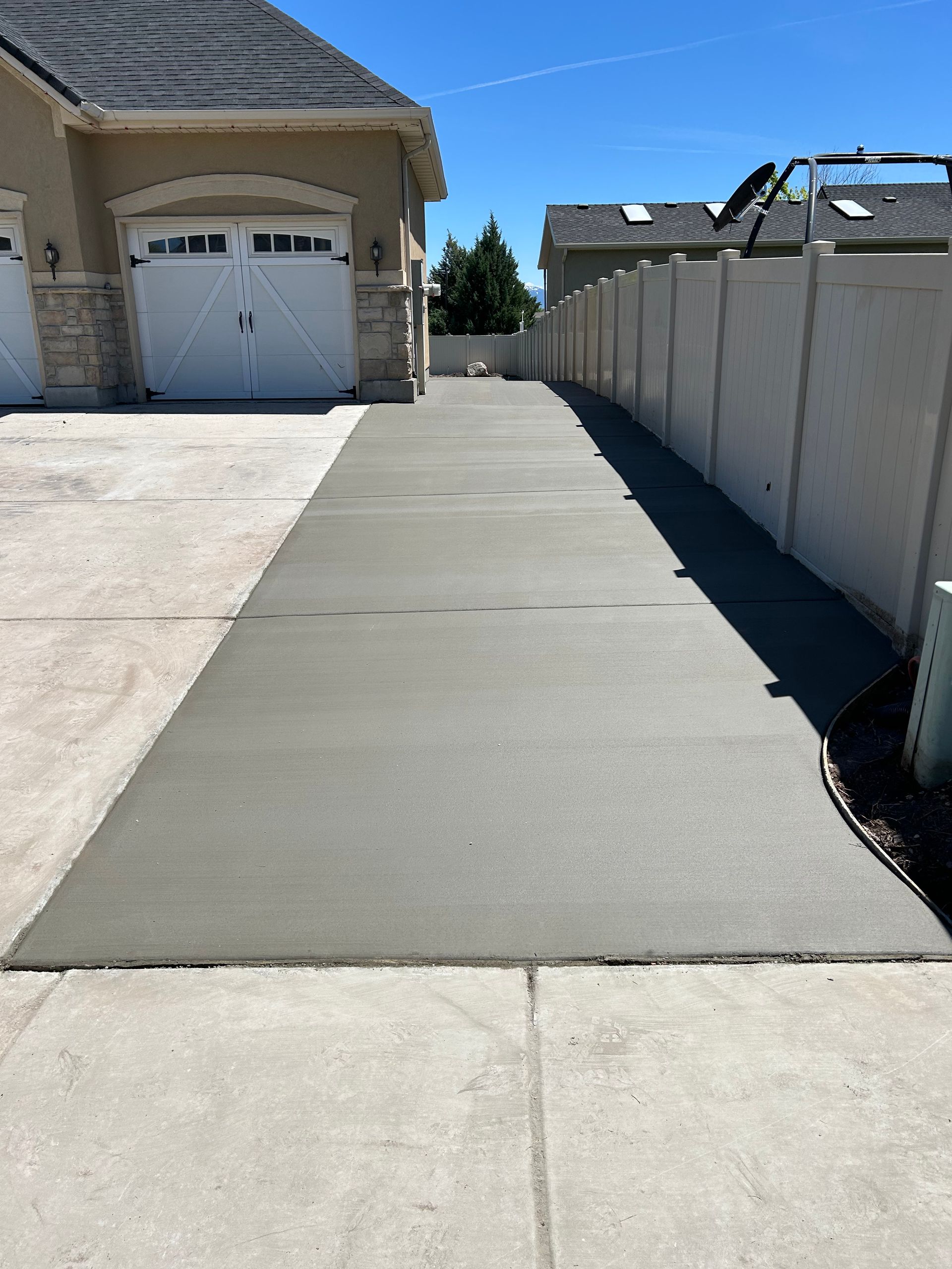 Concrete driveway and walkway beside a house and tall fence under bright sunlight