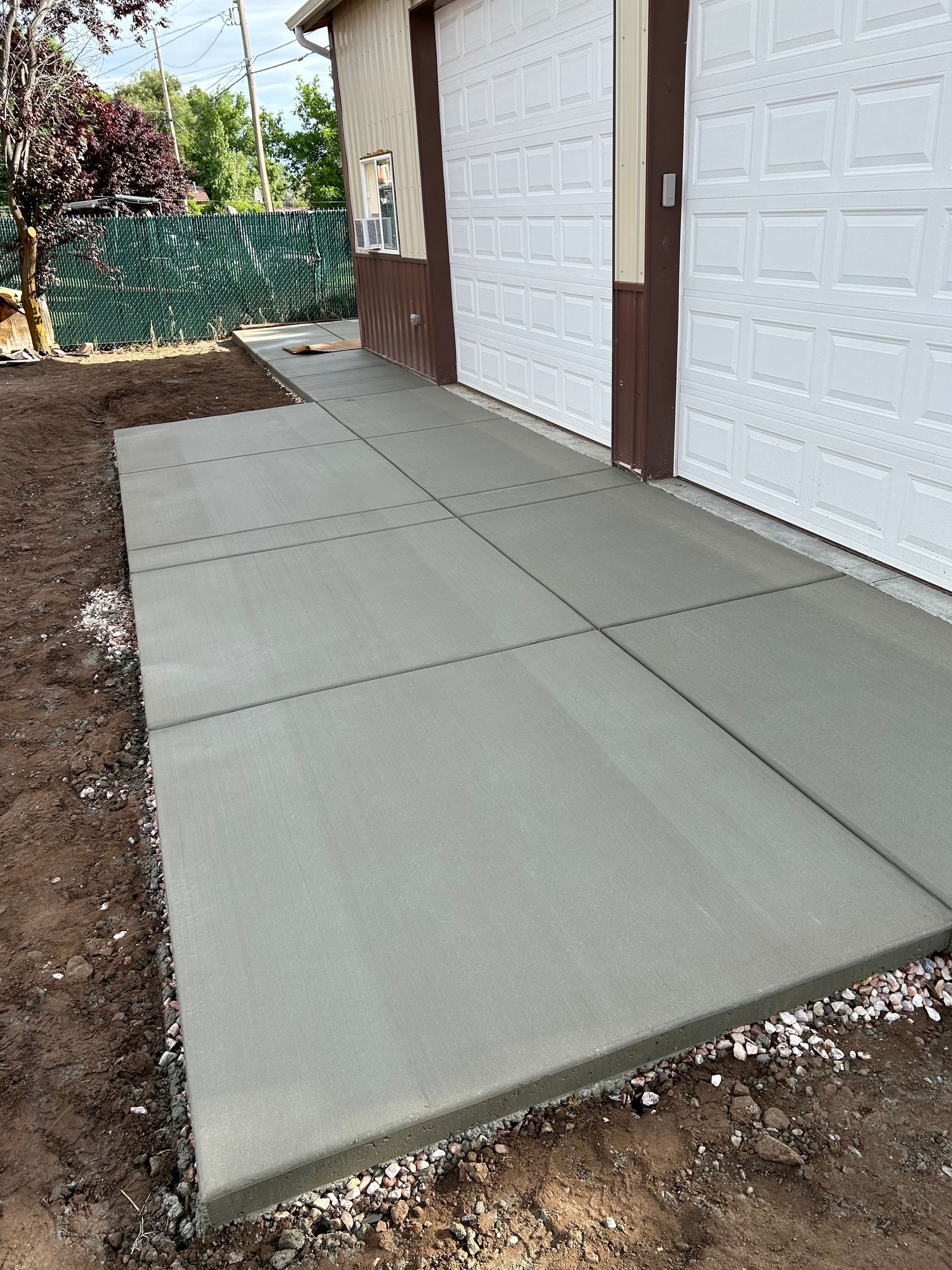 Fresh concrete walkway leading to white garage doors beside a dirt yard and fence