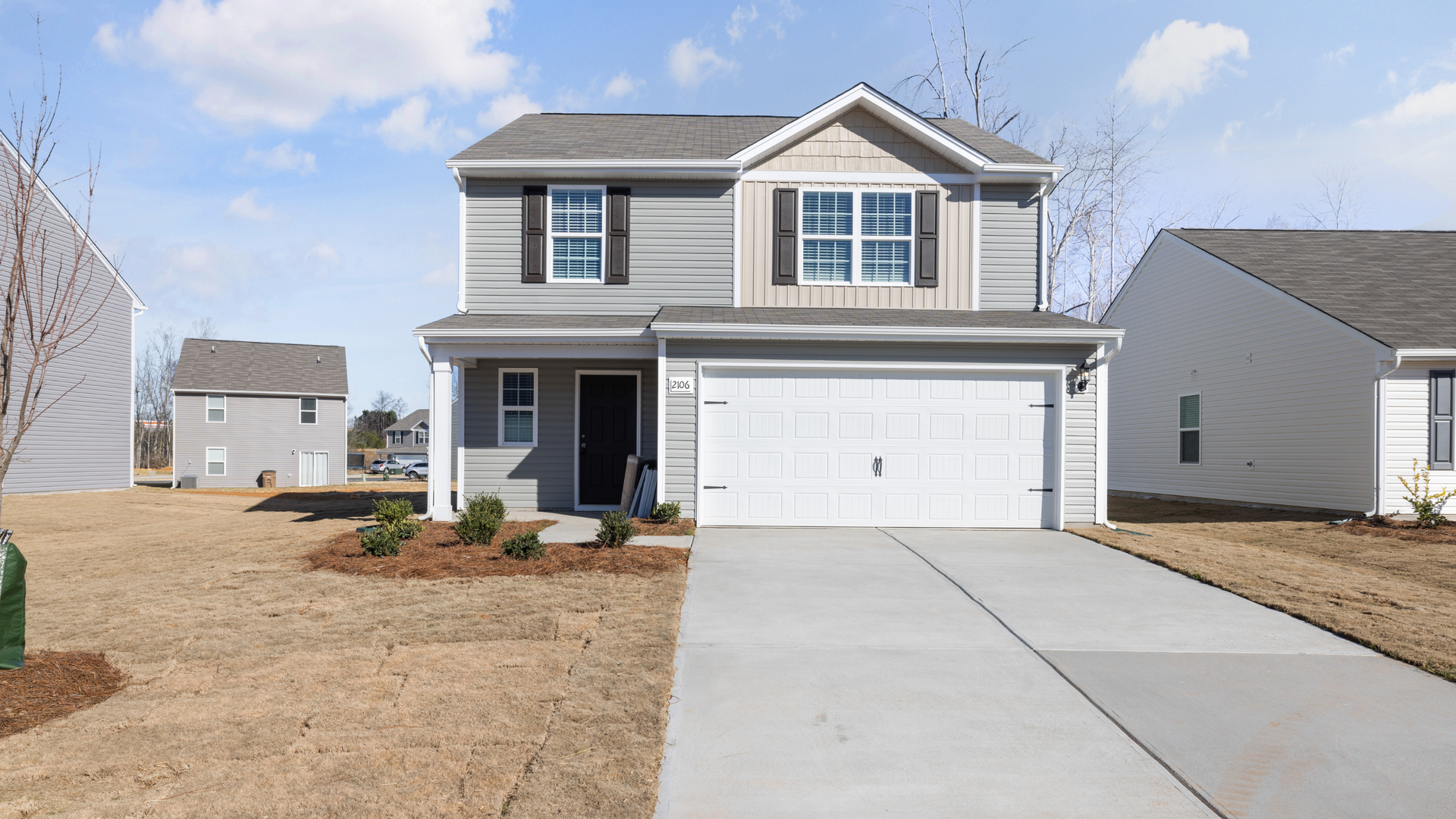 A two-story grey vinyl siding house with a white garage door, front porch, and concrete driveway under a blue sky.