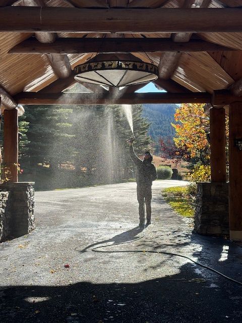 Person sprays water upwards under a wooden porch with stone pillars. Sunlight shines through the water.