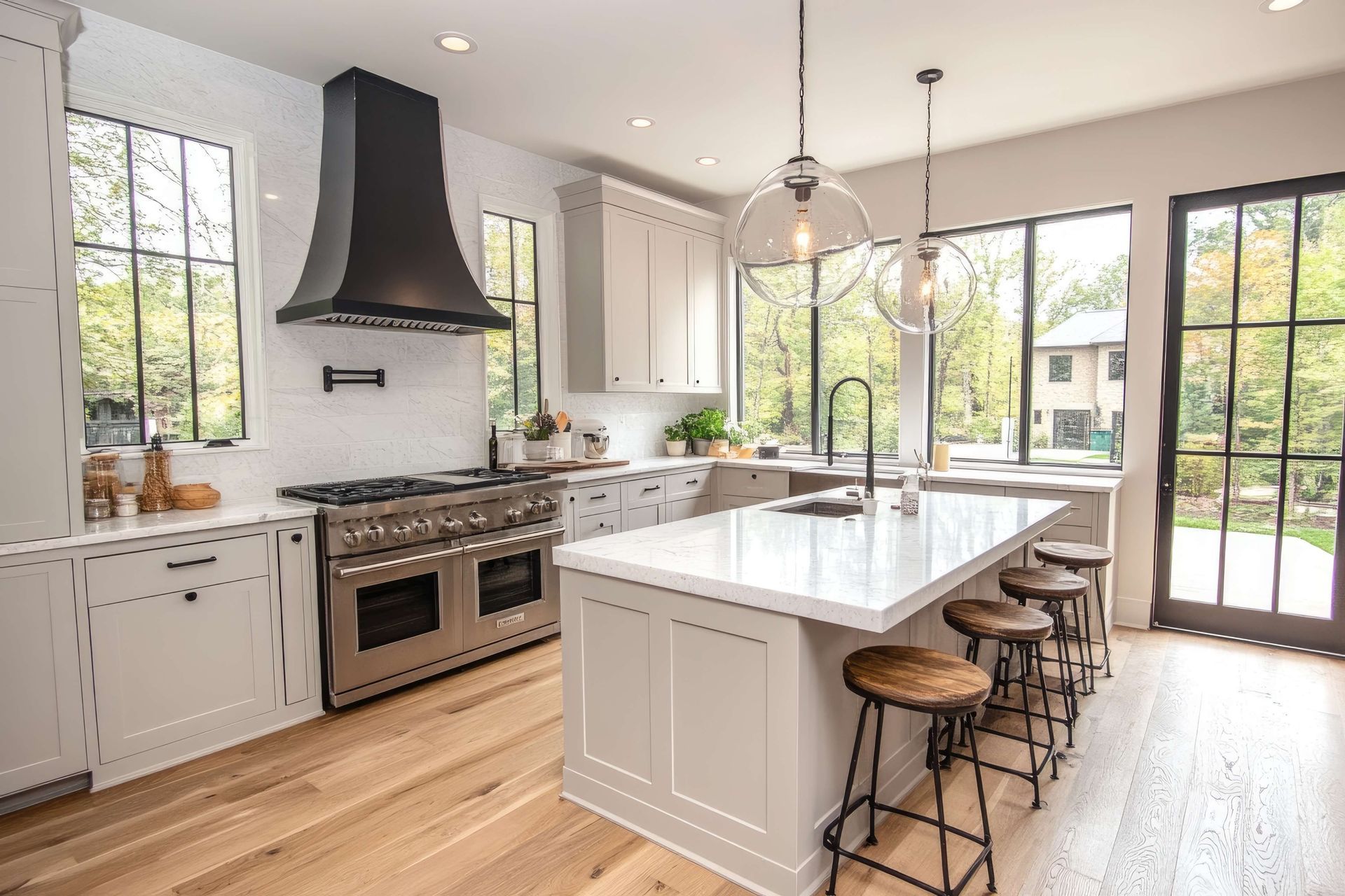 Bright modern kitchen with white cabinets, black range hood, large island, and tall windows overlooking trees