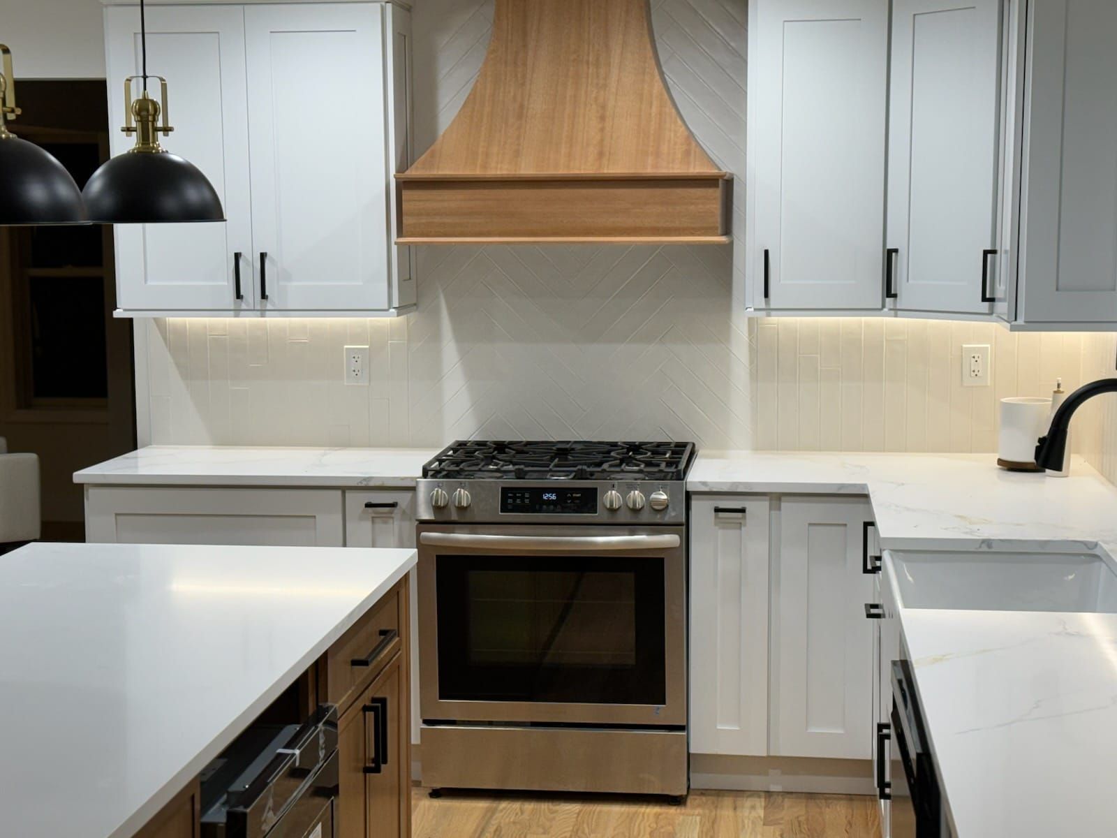 Modern white kitchen with stainless steel stove, wood range hood, and black pendant lights