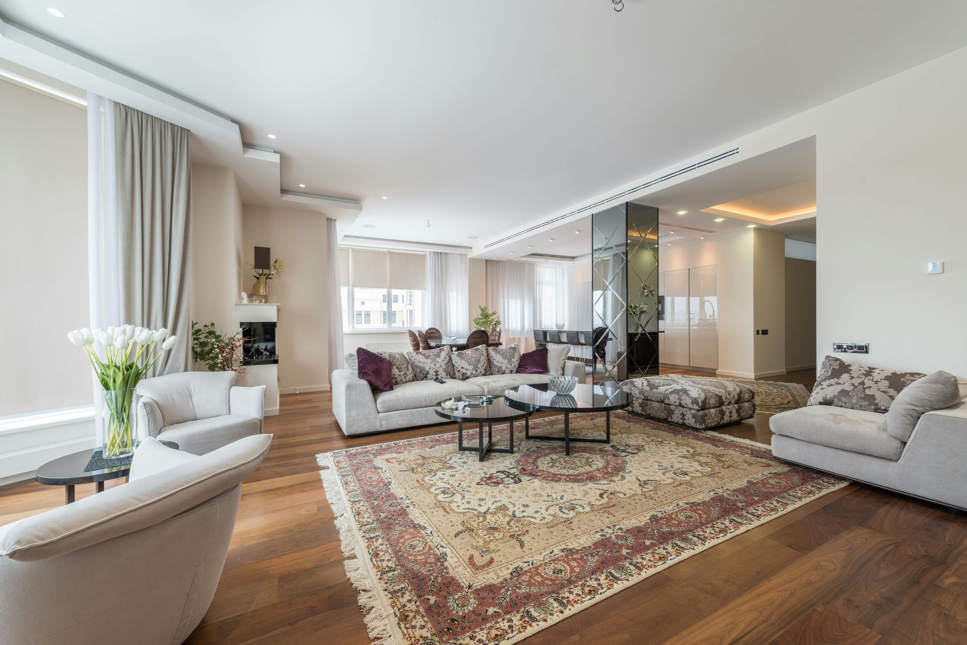 Bright modern living room with beige sofas, patterned rug, wood floor, and large windows